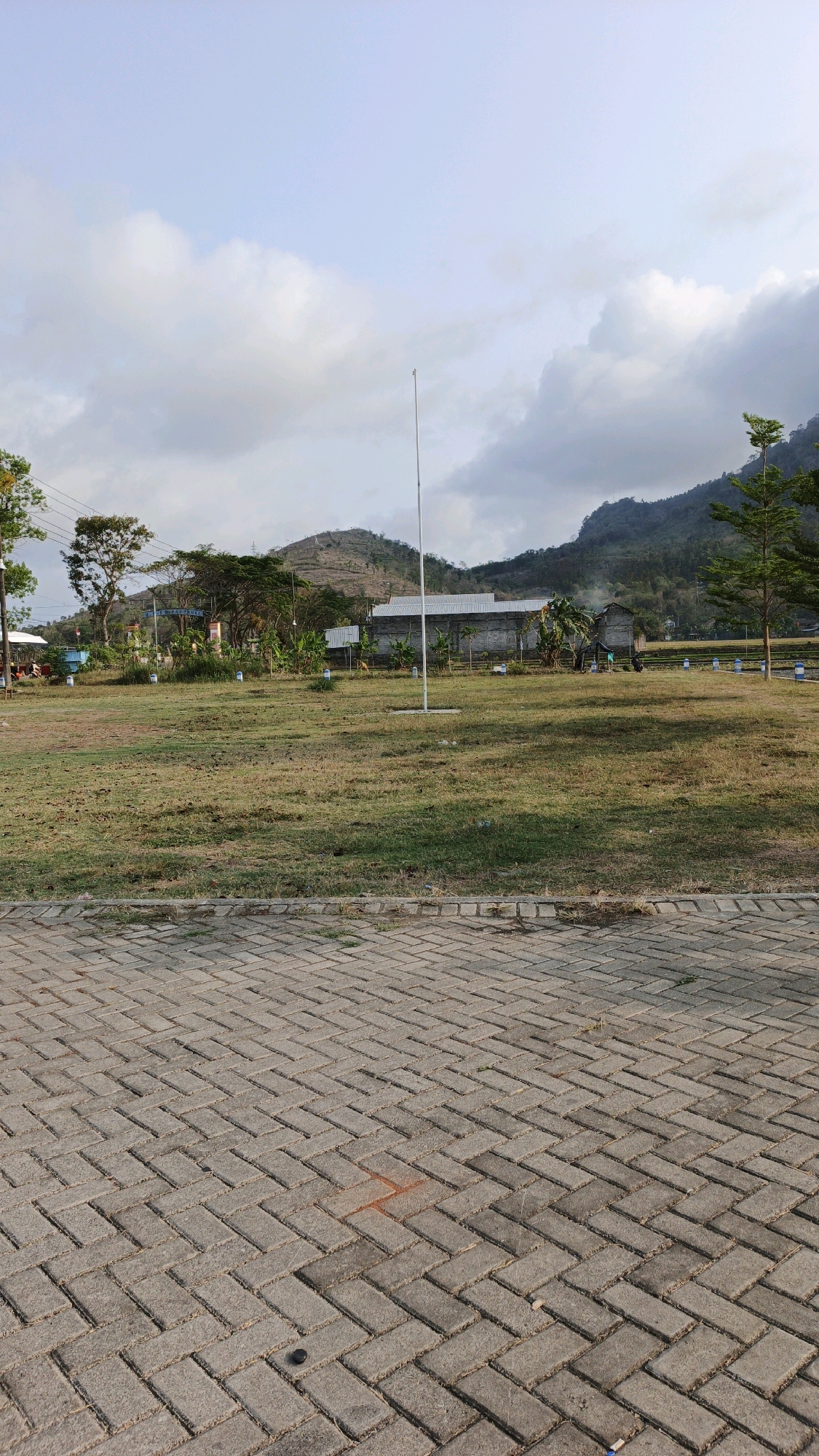The image depicts an outdoor scene in Tanggulwelahan, Indonesia. In the foreground, a herringbone-patterned brick walkway stretches across the bottom of the frame. Beyond the walkway, a large grassy field occupies the middle ground, with scattered patches of dry grass and some small bushes. A tall, thin flagpole stands centrally in the field. In the background, several low-lying buildings with corrugated metal roofs are visible, interspersed with trees. Behind the buildings, rolling hills covered in vegetation and some terraced areas form the backdrop, leading up to a mountain range under a partly cloudy sky. The overall impression is of a semi-rural or park-like setting with buildings and natural landscape elements.
