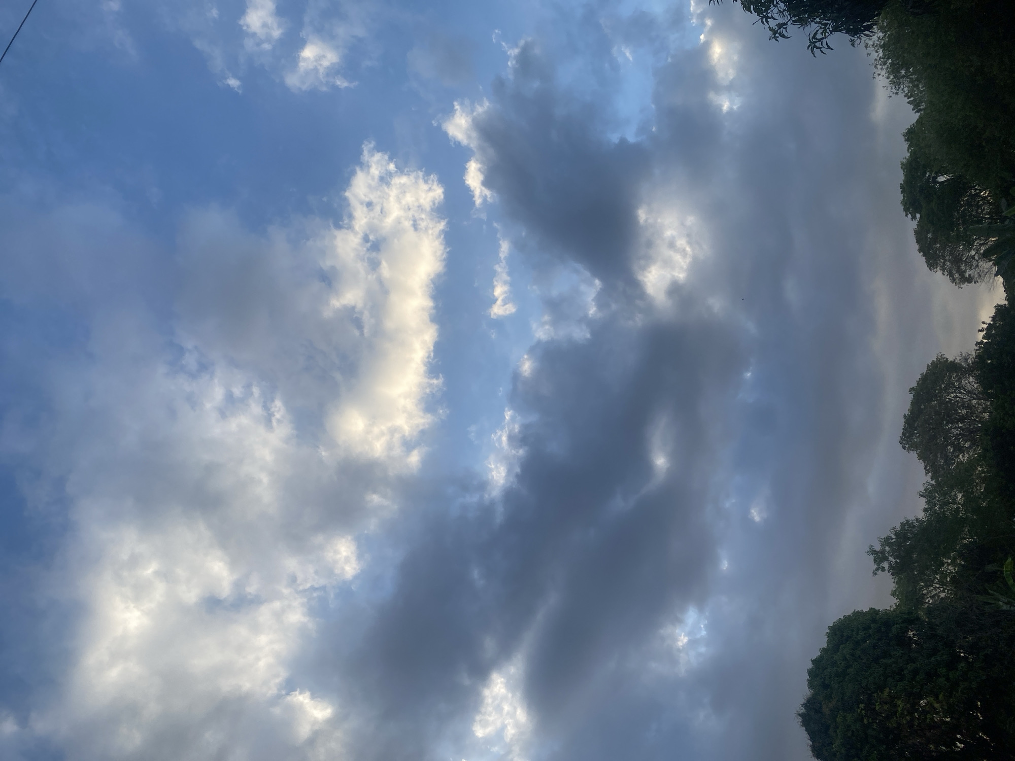 The image shows a sky filled with clouds in the city of Banjarbaru, Indonesia. The clouds are a mix of dark grey and bright white, with patches of blue sky visible. The lower portion of the image is dominated by the dark silhouettes of trees and foliage, suggesting a natural or semi-rural setting within the city. The lighting indicates it could be either sunrise or sunset, or an overcast day with breaks in the clouds. No people or specific man-made structures are prominently featured, focusing the scene on the natural elements of the sky and vegetation.