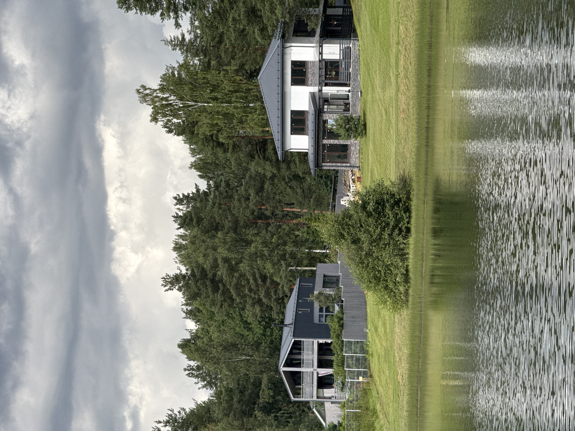 This image captures a waterfront scene with two modern houses situated behind a fence, backed by a dense forest. The sky is overcast with dramatic, dark clouds. In the foreground, a calm body of water reflects the surrounding greenery and the sky.
The house on the left is a dark gray, two-story structure with a distinctive angled roofline and prominent balconies. It appears to be constructed with a mix of materials, including wood or composite siding. A light-colored fence with vertical posts separates the property from the grassy embankment.
The house on the right is a lighter-colored, multi-story dwelling with large windows and a stone facade on the lower level. It features a dark-tiled roof and various architectural elements, including covered porch areas. A small evergreen tree stands in front of this house.
The environment is lush and green, with a well-maintained lawn leading down to the water's edge, where reeds and tall grass grow. The forest behind the houses is composed primarily of coniferous and deciduous trees, creating a natural backdrop. The overall impression is one of a serene, contemporary residential area bordering a natural landscape. There are no people or animals visible in the image. This location is not in the city of Mars, Russia.