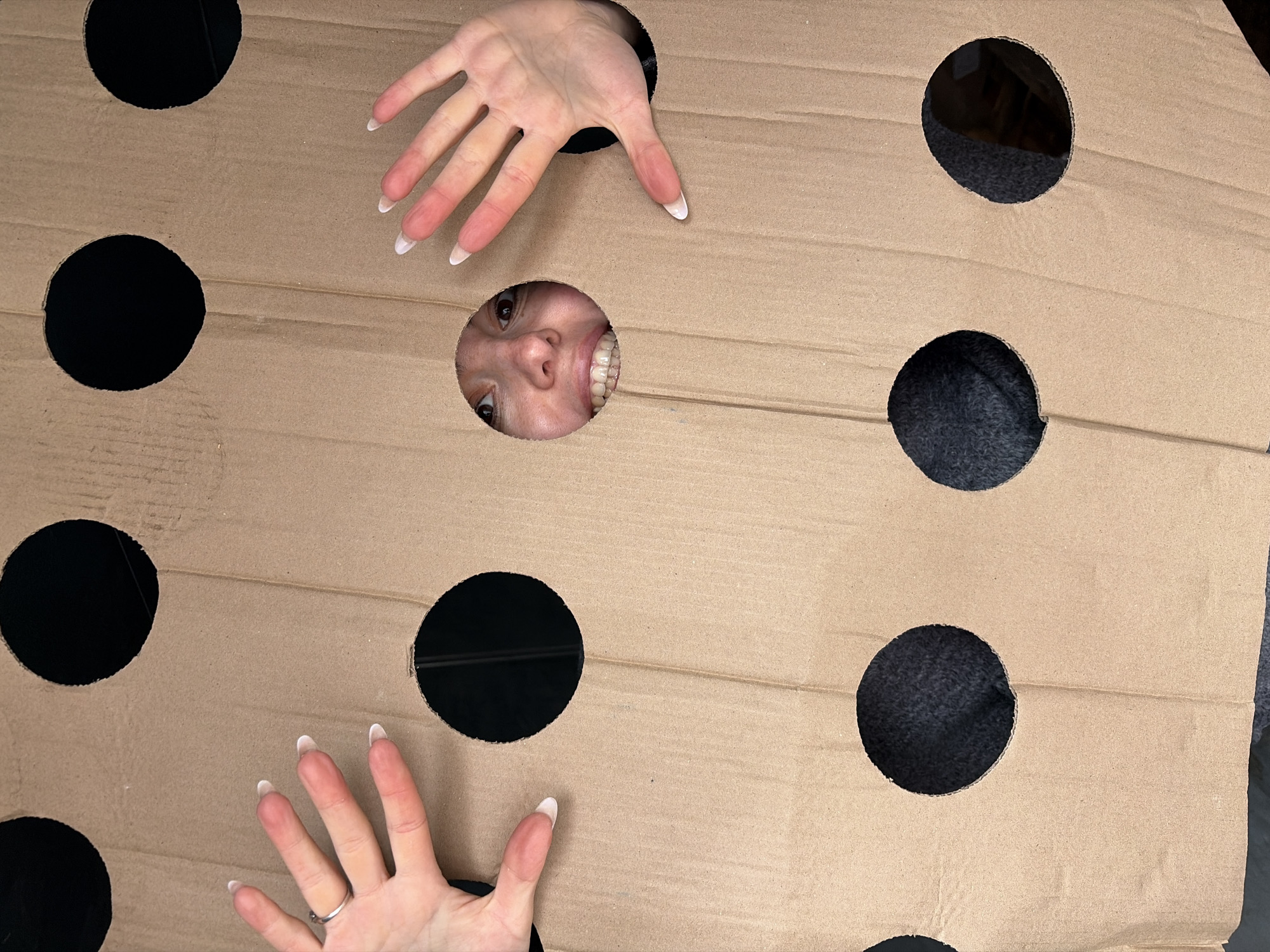 A woman's face and two hands emerge from a large piece of brown cardboard with multiple circular holes cut out. The woman's face is visible through a central hole, while her hands extend from holes above and below. The background seen through the remaining holes is dark, suggesting an enclosed space. The hands have manicured nails. The cardboard appears to be a makeshift game or prop. There is no indication of Izhevsk, Russia.