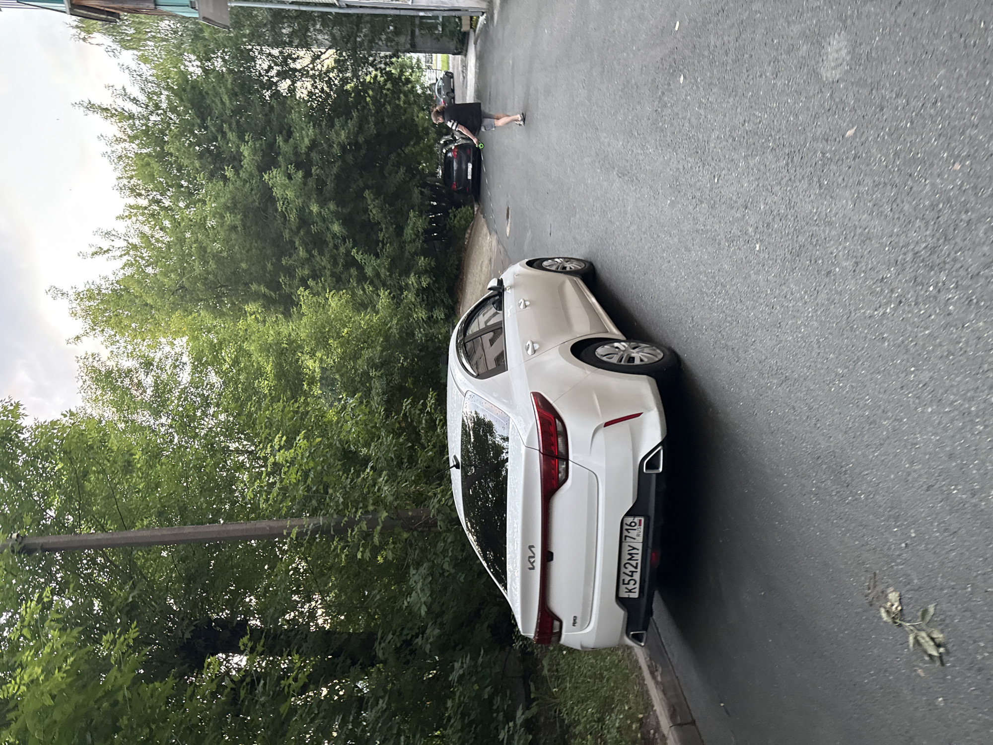 The image shows a street scene in Kazan, Russia. A white Kia Rio sedan is parked on the left side of the asphalt road, facing away from the viewer. The car has a license plate that reads "K542MY 716 RUS". A person with short hair, wearing a black vest over a white and black striped shirt, grey shorts, and blue sandals, is walking away from the car on the right side of the road. The person is holding a green object, possibly a leash or a toy. Several trees line the left side of the street, and a building with a light-colored facade is visible on the right side. Another dark-colored car is parked further down the street. The sky is overcast.