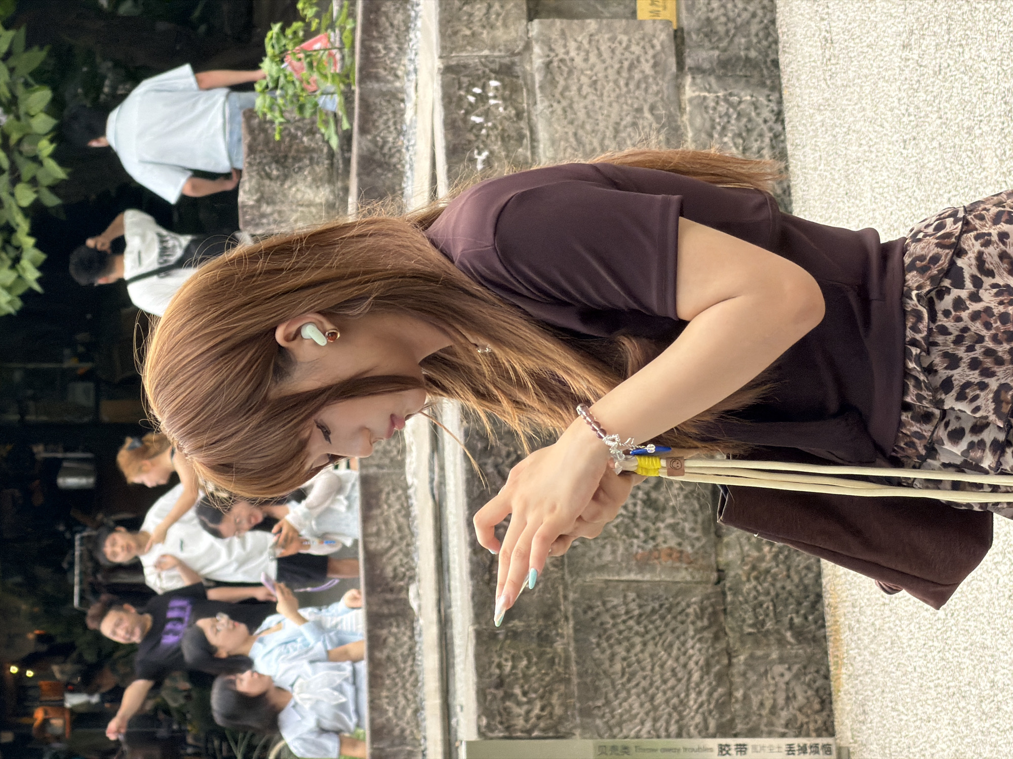In the Yuzhong District of China, a young woman with long, reddish-brown hair and a brown t-shirt is the central focus of the image. She is holding a smartphone in her hands and appears to be typing or scrolling. She has on light blue nail polish and is wearing a light blue earring. Her right arm is bent, and her left arm is resting on her bag. A brown handbag hangs from her left arm, with yellow straps and a beaded bracelet on her wrist. In the background, a group of people are visible. Some are standing, and one person appears to be pointing. The setting seems to be an outdoor area with a stone wall and some greenery.