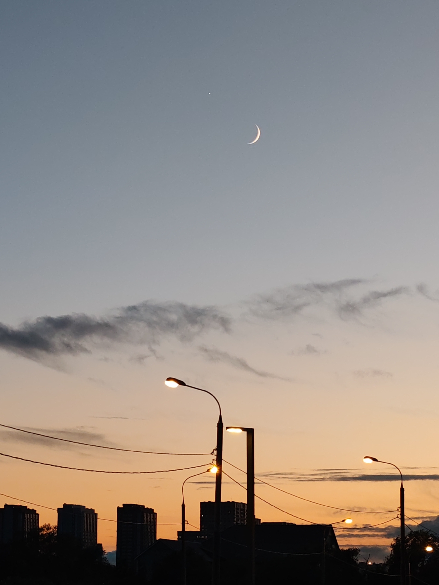 A crescent moon hangs in the pale blue sky, accompanied by a small, bright star. Below the celestial bodies, a band of dark clouds stretches horizontally across the frame. The lower portion of the image is dominated by the silhouette of a cityscape at dusk. Tall, modern buildings, some with sharp, angular rooftops, are visible. Several streetlights are illuminated, casting a warm glow on the scene. Power lines crisscross the sky, adding to the urban atmosphere. The overall impression is one of a city at twilight, with a hint of celestial beauty above.