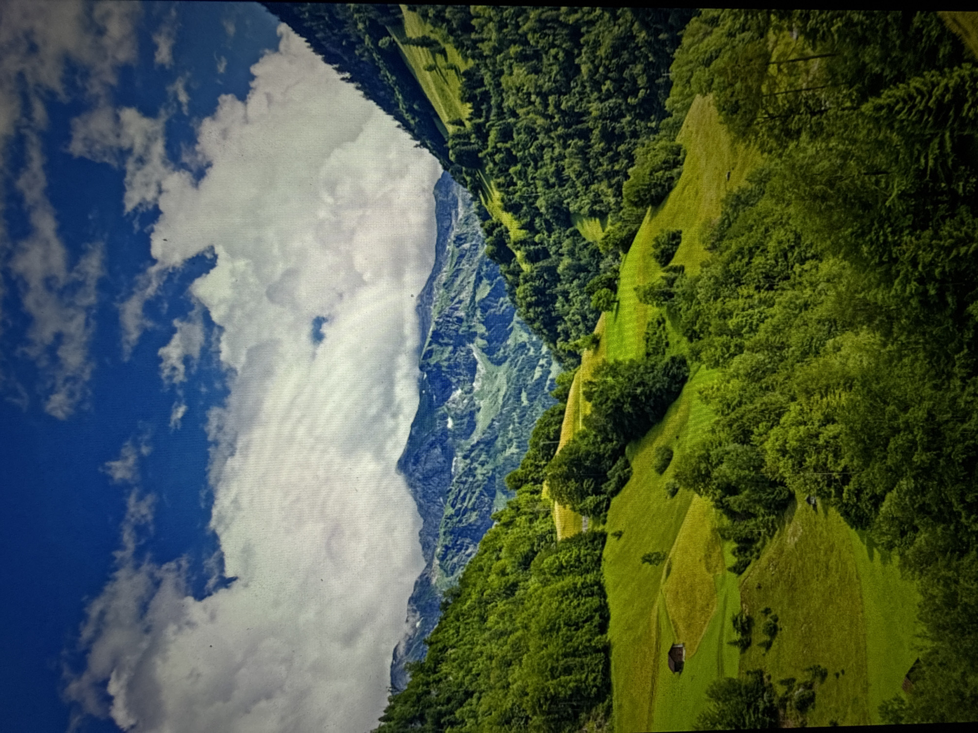 This is a vertical, high-angle shot of a lush green mountain landscape. The sky is a vibrant blue with dramatic white clouds. Rolling hills covered in vibrant green grass and dense forests fill the midground and foreground. In the distance, rugged mountains rise, some with patches of snow. Scattered throughout the landscape are small, dark brown wooden buildings, resembling cabins or barns, nestled into the hillsides. The overall impression is one of a serene and picturesque natural environment.