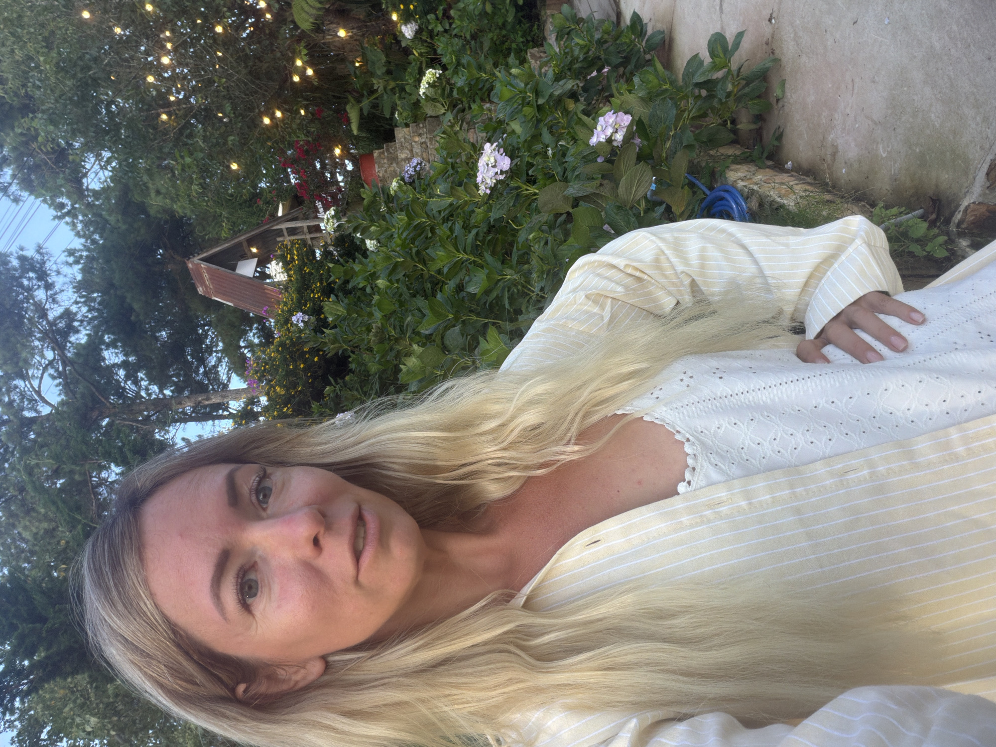 This is a vertical, selfie-style medium shot of a blonde woman lying outdoors. She is wearing a light yellow and white striped button-up shirt with a white eyelet camisole underneath. Her long, wavy blonde hair is spread out around her. She is looking directly at the camera with a neutral expression.
The setting appears to be a garden or yard. Behind her, there is lush green foliage with some pink and purple flowers. In the background, string lights are visible, suggesting it might be evening or late afternoon. A rustic wooden structure with a corrugated metal roof is also visible among the trees, along with some electrical wires. A blue hose is coiled on the ground near a stone wall. The lighting suggests it is daytime, possibly late afternoon or early evening, with warm, natural light.
The woman's location is contextually identified as Xuân Thành, Vietnam. There is no visible text in the image. The overall mood is relaxed and serene.