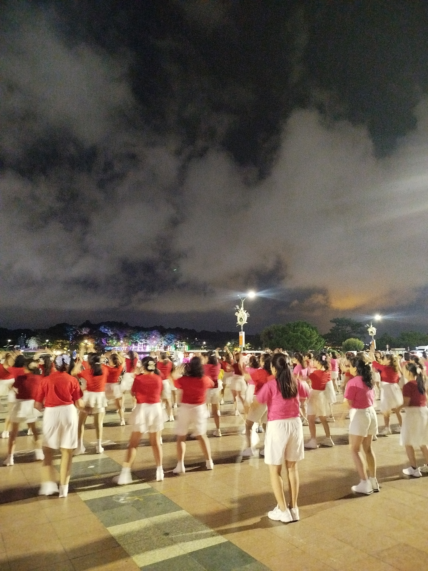 A large group of young women, seen from behind, are performing a dance routine outdoors at night. They are all wearing matching pink tops and white shorts or skirts, and white shoes. The women are arranged in rows, facing away from the camera, with some in focus in the foreground and others further back. The ground they are dancing on appears to be a paved plaza with patterned tiles. In the background, there are trees lit with colorful lights, and streetlights are visible, indicating an urban or park setting. The sky is dark and cloudy, suggesting it might be nighttime or overcast, with some faint light suggesting the presence of artificial lights or a distant glow. The overall scene appears to be a lively performance or synchronized activity taking place in a public space.