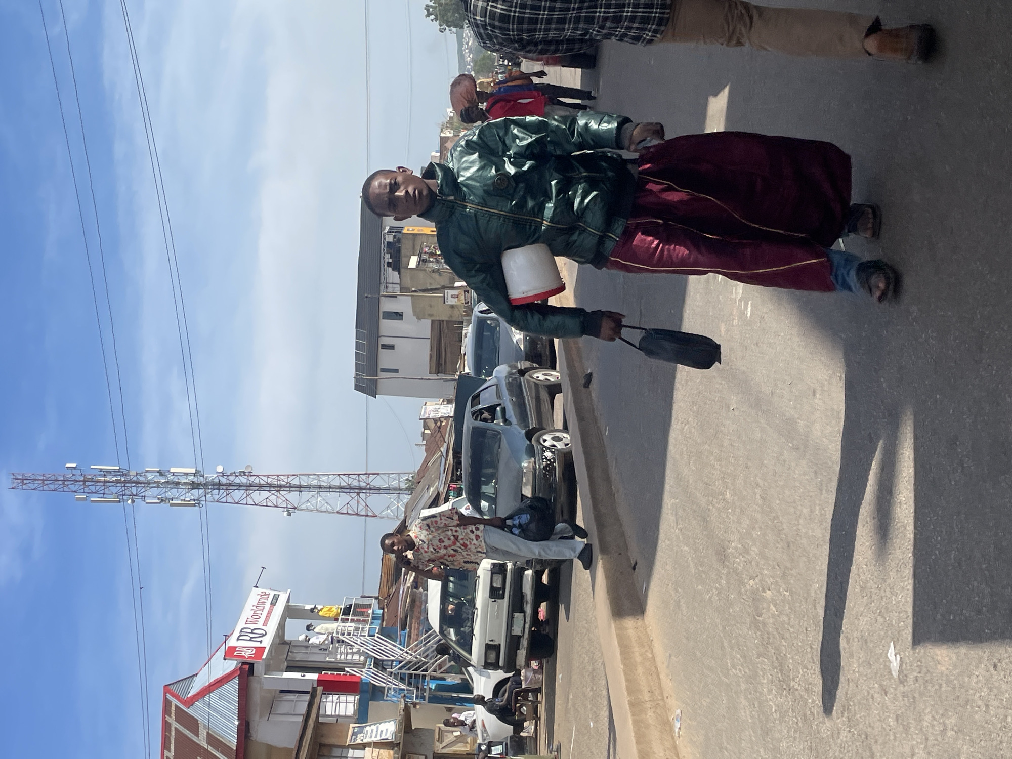 The image depicts a street scene in Jos, Nigeria, dominated by a tall telecommunications tower. In the foreground, a young man wearing a dark green jacket and a maroon garment, possibly a wrapper, walks towards the camera. He is carrying a white bucket and a black bag. Behind him, another man in a patterned shirt walks away, carrying a black bag. To the left, a white van and a dark-colored sedan are parked on the side of the road, with a man sitting on a stool near the van. Several buildings line the street, including one with a prominent "RB Worldwide" sign. The sky is clear and blue with some scattered clouds. The ground is paved, and a curb runs along the side of the road. The overall impression is of a busy urban environment with pedestrian and vehicular traffic.