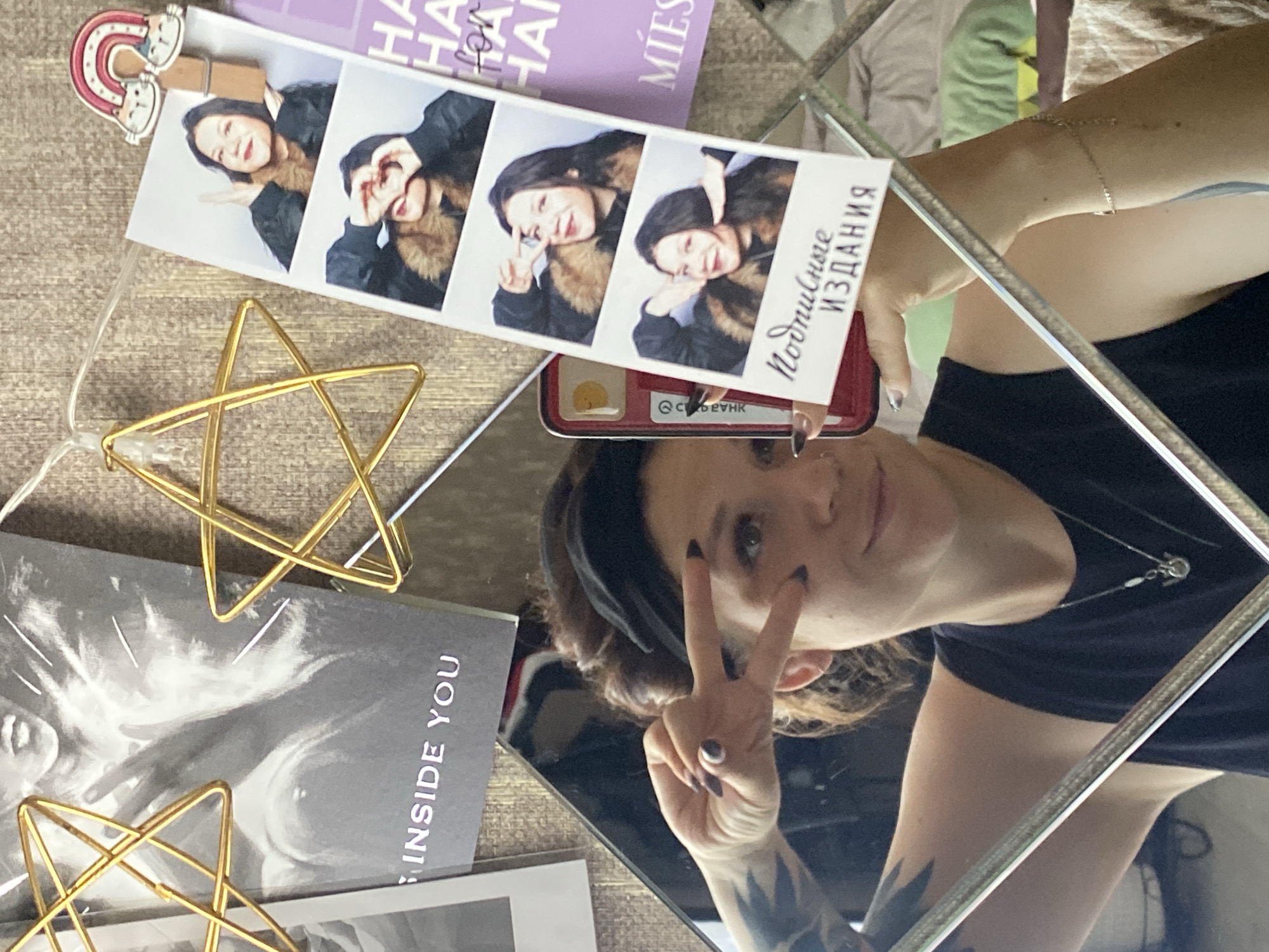 This is a close-up, eye-level, indoor, medium shot of a woman taking a selfie in a mirror. The woman, who appears to be in her late 20s or early 30s, has dark hair pulled back in a bun and is wearing a black tank top and a black headband. She is making a peace sign with her right hand, her fingers are painted with dark nail polish. Her left arm is visible in the mirror, showing a tattoo on her upper arm and a delicate bracelet on her wrist. A necklace with a small pendant is also visible. The background consists of a wall decorated with various items, including two gold wire star ornaments, a black and white poster with text that reads "INSIDE YOU", a strip of photo booth pictures of the woman, and a purple card with text that includes "HA HA" and "MIES". The photo booth strip shows three images of the woman posing, with text at the bottom that appears to be in Russian. The overall environment suggests a personal space, possibly a bedroom or a living area. There is no specific information in the image that can identify the location within the city of Chaikovsky, Russia.
