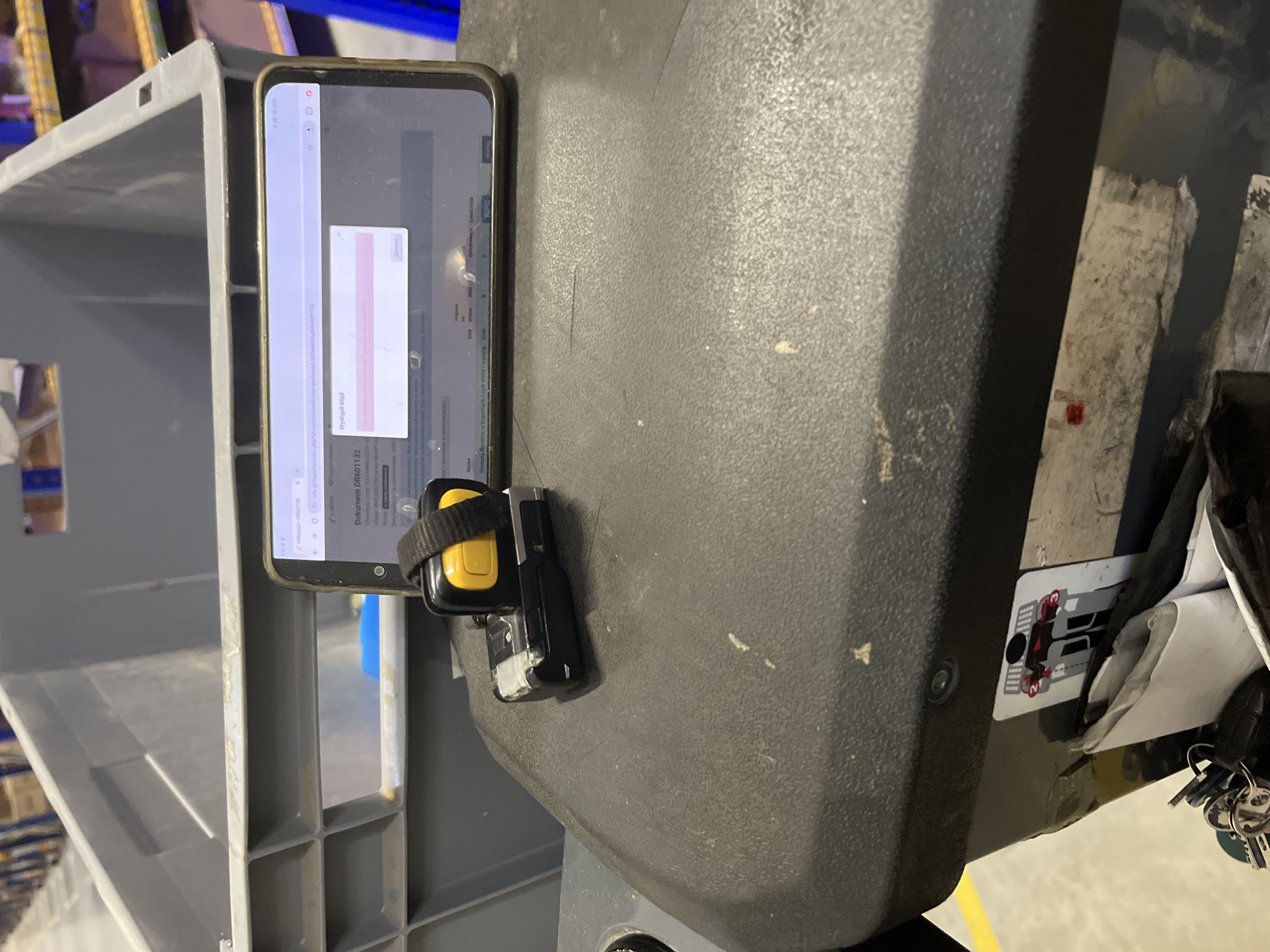 The image depicts a close-up view of a black textured surface, likely the dashboard of a vehicle or a piece of industrial equipment, in Tarnowo Podgórne, Poland. A smartphone is propped up, displaying a webpage with Polish text. A small device with a yellow button and a black strap is attached to the smartphone. The background shows large grey plastic bins, commonly used in warehouses or logistics facilities. There are also some keys and what appears to be a white paper object visible in the lower part of the image. The scene suggests a work environment, possibly related to inventory management or logistics.
