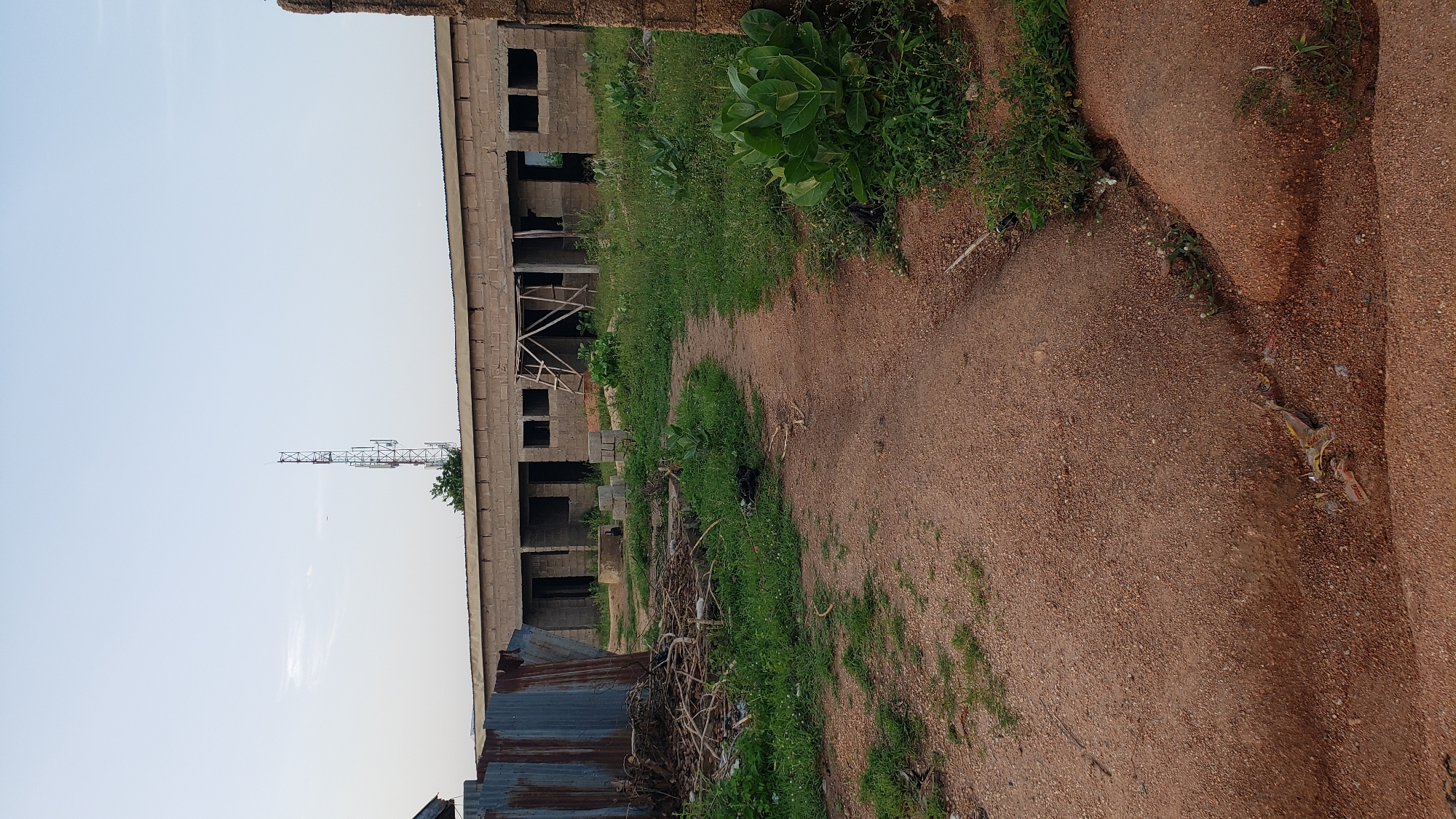 The image depicts an outdoor scene in Jauro Yinu, Nigeria, characterized by a partially constructed building and its surroundings. The building, made of concrete blocks, appears to be a long, single-story structure with open doorways and window openings. Scaffolding is visible against one section of the building. In the foreground, a dirt path winds through overgrown green grass and vegetation. To the left of the path, there is a pile of debris including what appears to be scrap metal sheeting and branches. Further in the distance, a tall telecommunications tower stands against a pale sky. The ground is reddish-brown soil, with some scattered litter. The overall impression is one of an undeveloped or in-progress construction site. There are no people visible in the image.
