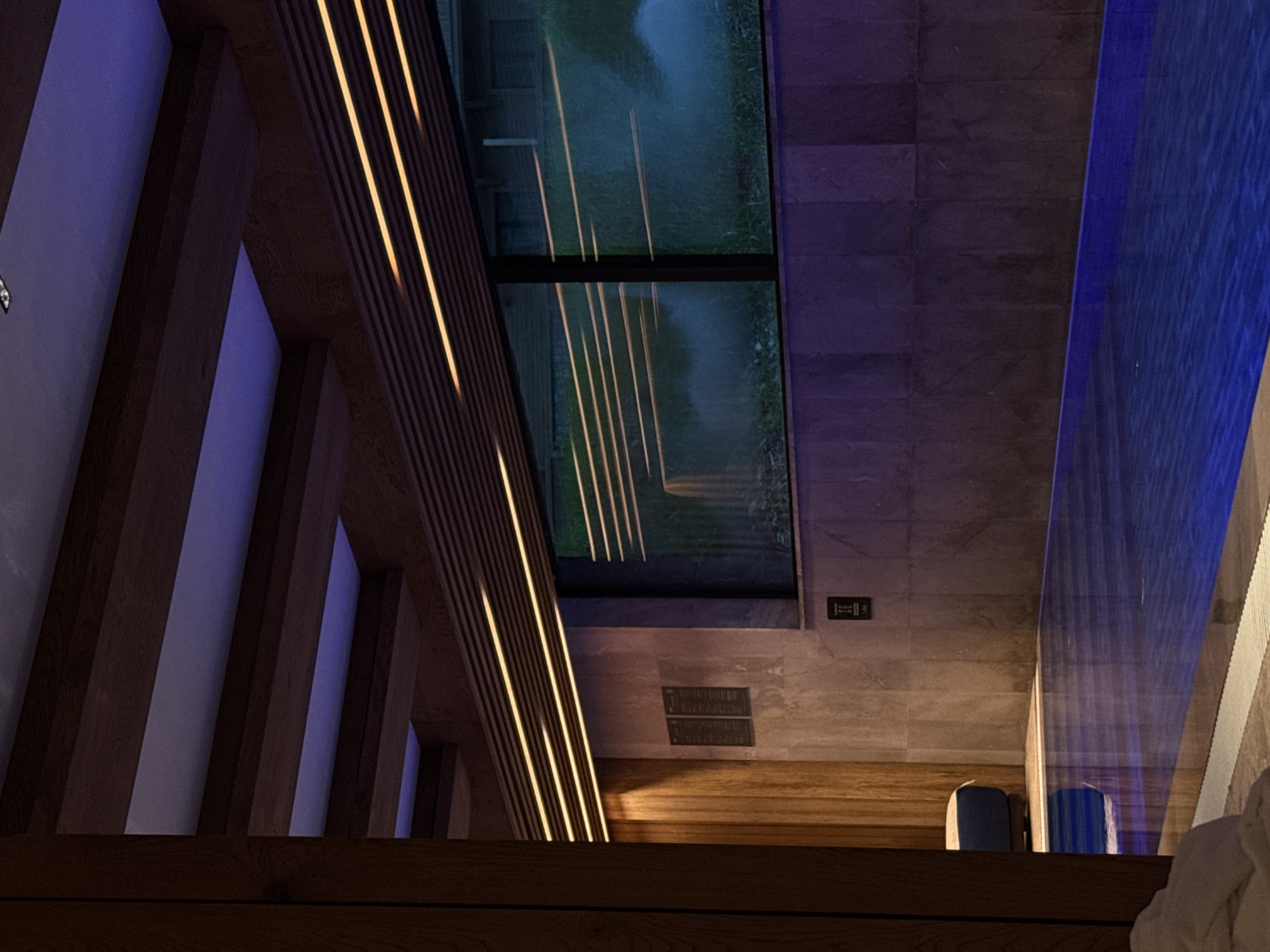The image depicts an interior view with a modern design aesthetic. On the left, a structure with wooden beams and illuminated strips is visible. The light fixtures appear to be integrated into the wooden structure, emitting a warm glow.
A window, possibly a skylight, shows an outdoor scene with greenery, suggesting a natural setting. The window frame is black, and the exterior is slightly blurred.
The walls are a neutral gray with a textured surface. There are also elements that suggest a bathroom setting, including what appears to be a partially visible shower or bath area on the right with blue lighting. Two cylindrical objects, possibly containers or decorations, are near the base of the frame.
The image does not provide sufficient information to confirm its location in Katun Bačko brdo, Montenegro. There are no people visible in the image, and no interactions or actions are depicted.