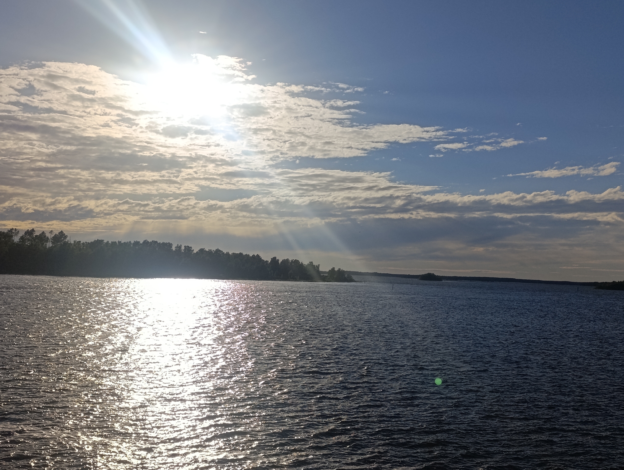 A wide shot shows a body of water reflecting the sun. The sun is bright, shining down through white, scattered clouds in a blue sky. The water is dark, with a bright reflection of the sun on its surface. Trees line the distant shore, silhouetted against the sky. The image appears to be taken from a boat or the shore, looking out over the water towards the horizon. It is a peaceful, serene scene. Based on visual cues such as the type of trees, the lighting and the expansive body of water, this scene is likely located in the city of Jakobstad, Finland, possibly on the archipelago.