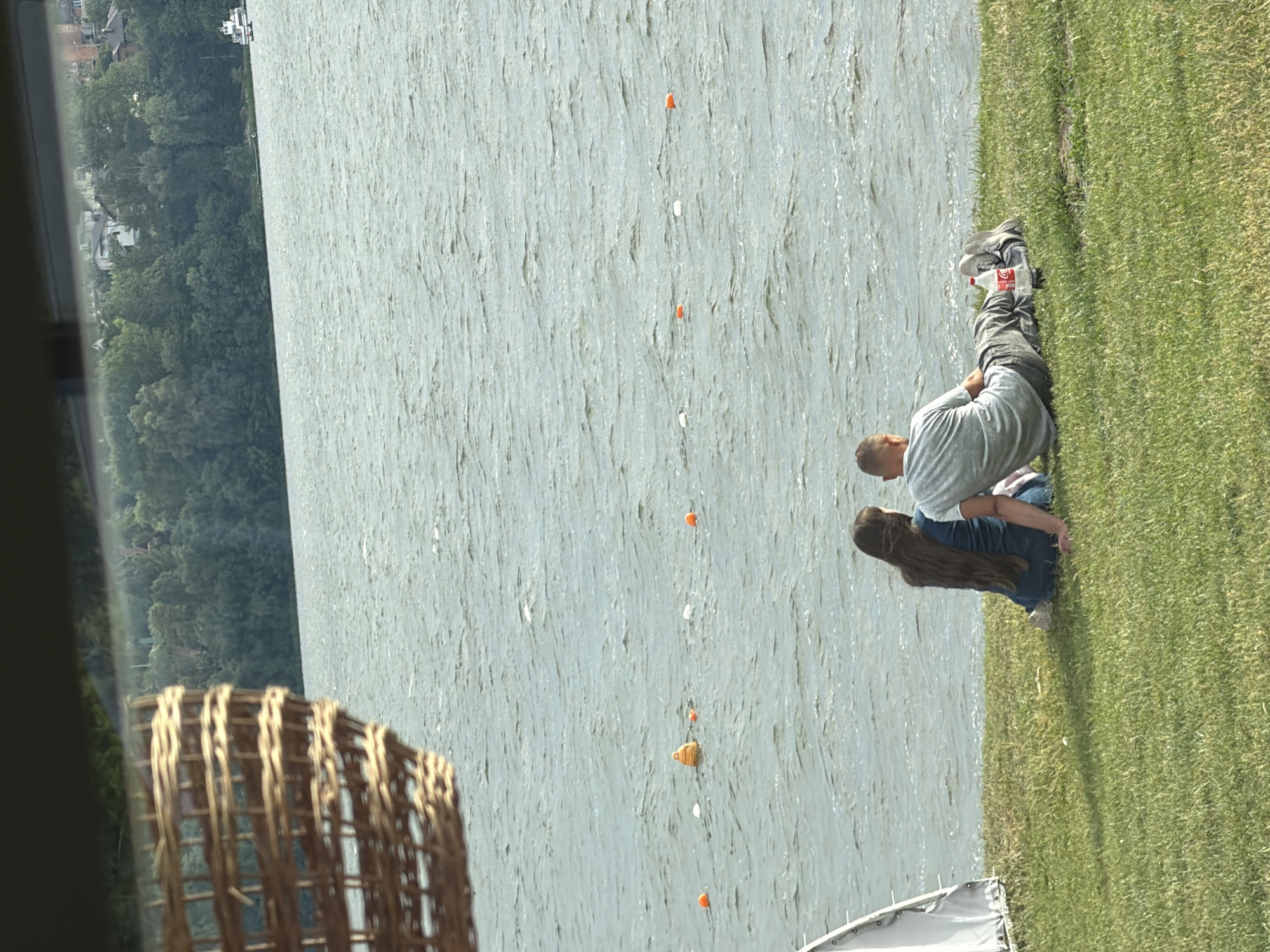 The image depicts a lakeside scene with a couple sitting on the grass, facing a body of water. The woman, on the left, has long brown hair and is wearing a blue top and jeans. The man, on the right, is wearing a grey t-shirt and jeans, and has a bottle of Coca-Cola placed on the grass next to him. In the background, the lake stretches out, with buoys visible on the water. The far shore is lined with trees and buildings, suggesting an urban or semi-urban environment. A white tent-like structure is partially visible on the left side of the image. A woven basket is in the upper left corner, partially obscuring the view.
The image does not provide any explicit information that can definitively place it in Ternopil, Ukraine.