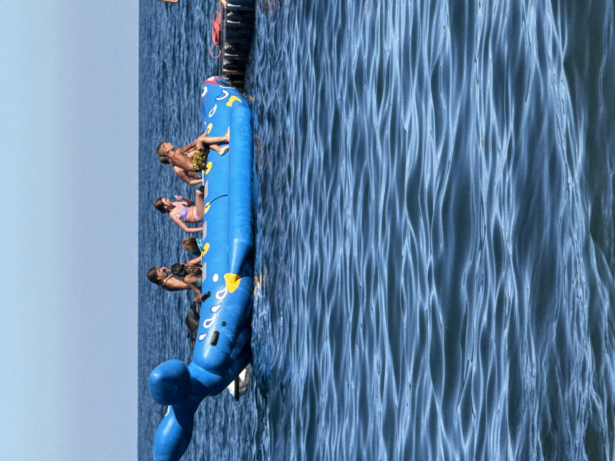 In Partenit, Ukraine, on a clear day, several children and a woman are enjoying a ride on a blue inflatable toy shaped like a whale. The toy is floating on the calm, blue sea, with gentle waves creating ripples on the water's surface. The children are seated on the whale, some are looking around, and one child is eating an ice cream. To the right of the whale, a dark-colored dock is visible, with a red rope coiled on it. The sky above is a clear, bright blue, indicating fair weather.