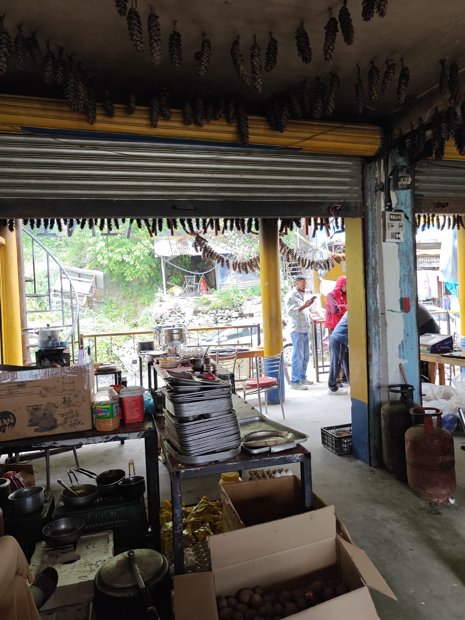 This vertical shot captures the rustic interior of an open-fronted eatery or kitchen in Pulga, India, offering a view of a lush green, hilly landscape. The ceiling is distinctively decorated with numerous dried pinecones hanging down, above a partially open rolling shutter.
In the midground, several people are present. A man in a light blue shirt and jeans stands, engrossed in his mobile phone. Another individual wears a red hooded jacket, and a third person in dark clothing is partially visible. Their presence suggests a casual gathering or waiting area.
The foreground reveals a functional kitchen setup. On the left, various cooking pots, pans, and utensils are arranged on low tables and a small, charcoal-burning stove. A large cardboard box with the text "2 Minute Noodles" and visible Hindi script ("बनाने..."), likely referring to Maggi noodles, is also present. On the right, stacks of empty metal trays are piled high on a table. Two red gas cylinders are positioned near a blue and yellow painted pillar. An open cardboard box filled with potatoes and another with eggs are visible on the floor, suggesting ingredients for cooking.
Beyond the eatery, the background features a vibrant green hillside covered in trees, with a spiral metal staircase and some rudimentary structures blending into the natural environment. A metal railing separates the indoor space from this scenic view. The bright lighting indicates it is daytime. On the far right, a blurry white sign has the partial text "KHEERGA".
