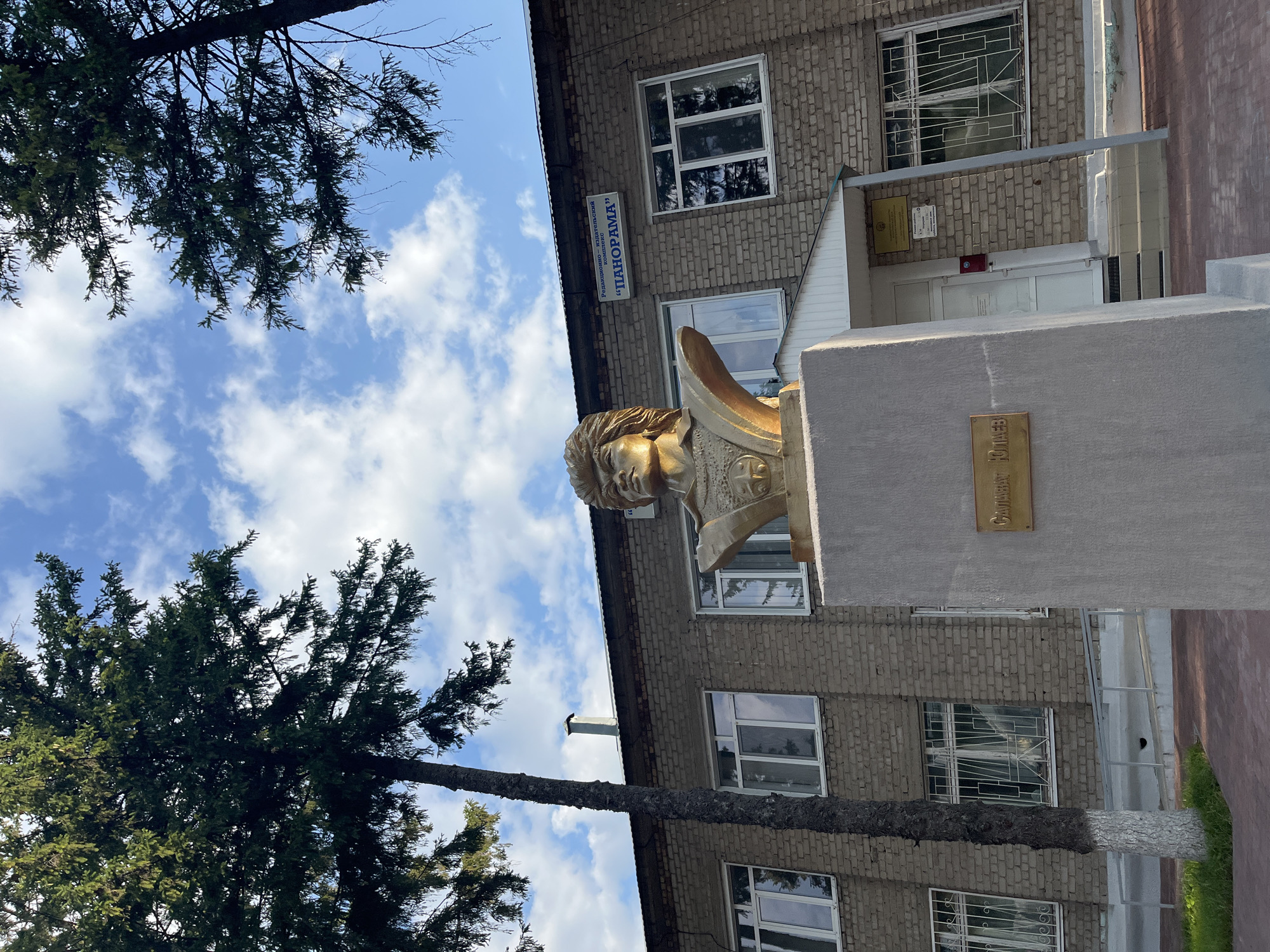 The image shows a bronze bust of a man on a concrete pedestal in front of a brick building. The bust is gold-colored and depicts a man with wavy hair. A plaque on the pedestal reads "САЛАВАТ ЮЛАЕВ" in Russian. The building has multiple windows, some with protective bars. A sign above a doorway reads "Редакционно-издательский комплекс «ПАНОРАМА»" in Russian. Two large evergreen trees frame the scene. The sky is blue with scattered white clouds. This monument is located in Blagoveshchensk, Russia, in front of the editorial and publishing complex "Panorama".