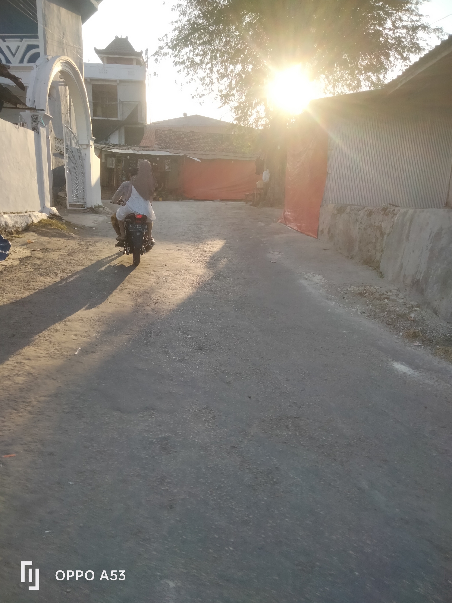 The image shows a dirt road in what appears to be a rural or suburban area. The sun is setting or rising, casting long shadows across the road and creating a lens flare effect. There is a motorcycle with two occupants, a driver and a passenger, riding away from the camera. The passenger is wearing a headscarf. To the left of the road, there is a white building with an arched entryway and an ornate gate. Behind the motorcycle, there are some buildings with corrugated metal roofs and orange tarps. The area appears to be under development or undergoing construction. The license plate on the motorcycle is visible and appears to be Indonesian. The watermarks at the bottom left indicate that the photo was taken with an OPPO A53 phone. Without further context, it is not possible to definitively pinpoint the exact location in Badung, Indonesia, but the scene is consistent with many areas in the region.