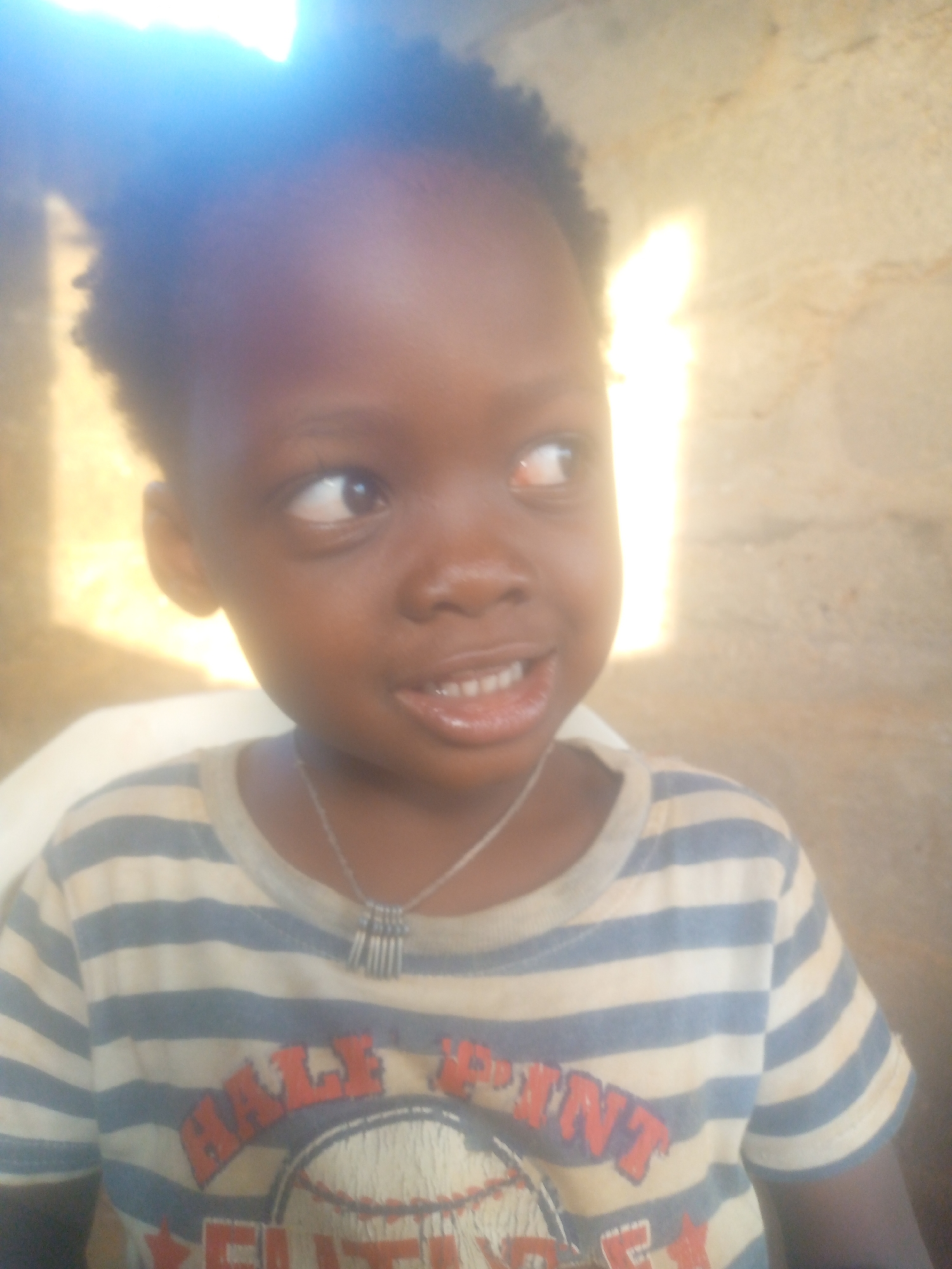 A young child with dark skin, short curly hair, and wearing a striped shirt and a necklace is looking to the right of the frame. The background is indistinct but appears to be a wall or other surface, illuminated by bright light. The location is unknown.