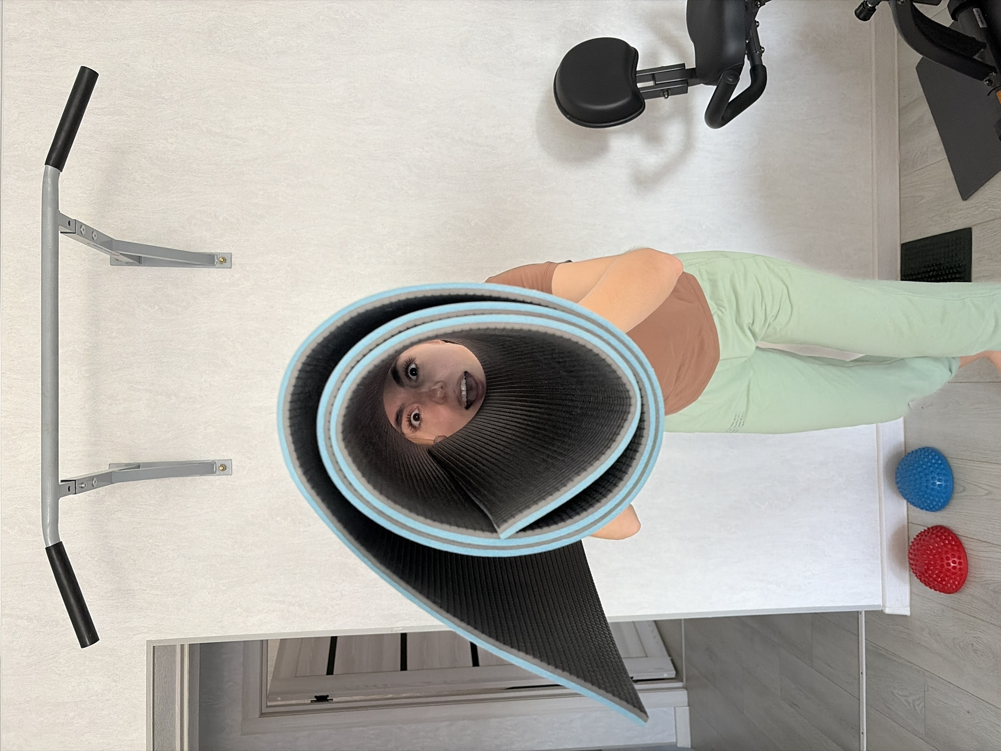 A young woman with fair skin and brown hair peers through the rolled-up end of a light-blue and black yoga mat. She wears a brown short-sleeved shirt and light-green pants. The mat is on a white padded bench, with a blue spiky massage ball and a red spiky massage ball on the floor nearby. In the background are gym equipment: a pull-up bar on the wall and a black massage chair. A window is visible near the bottom of the frame. The location is likely a home gym in Karyakino, Russia.