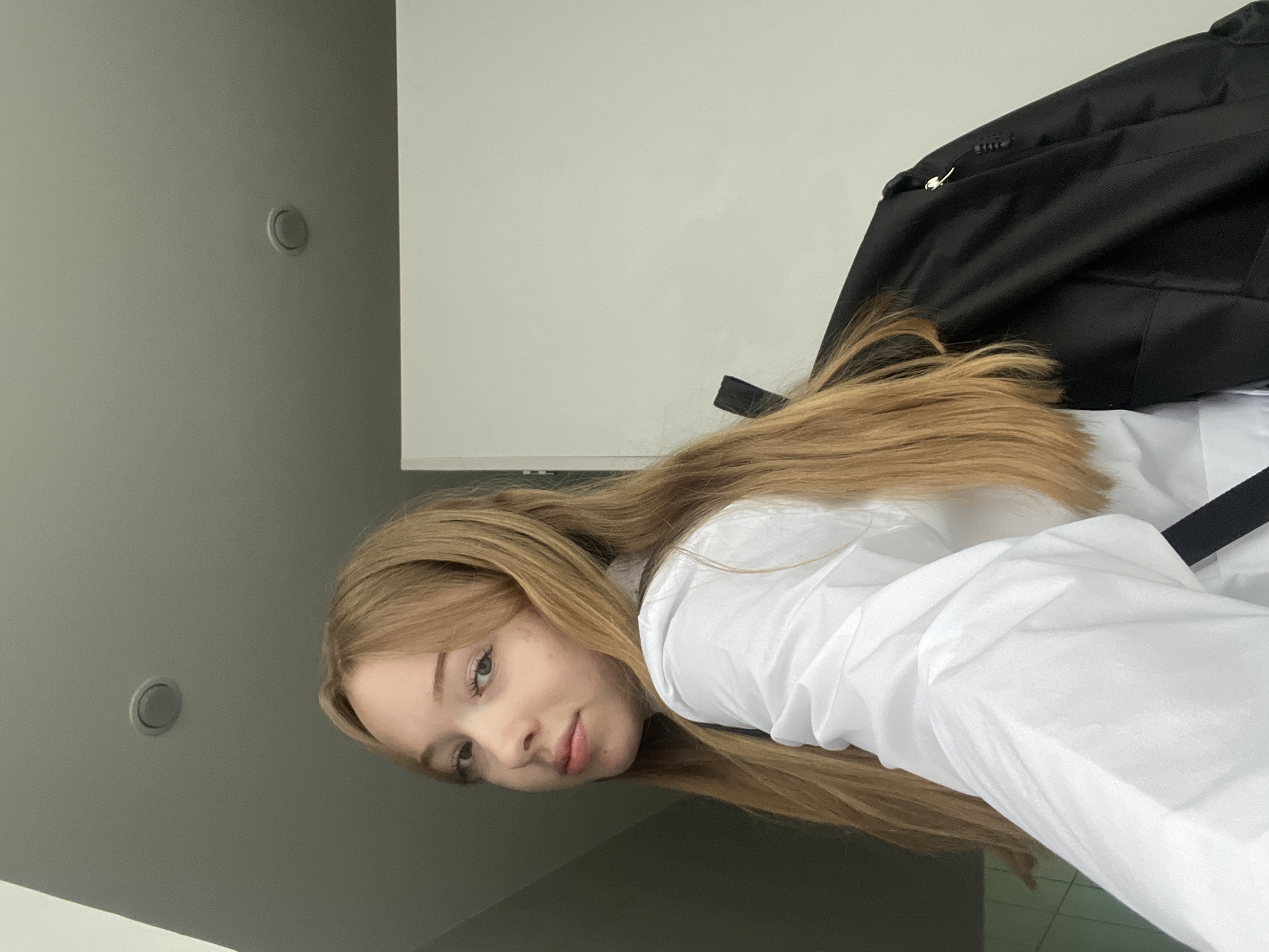 A young woman with long, blonde hair is looking directly at the camera. She is wearing a white collared shirt and a black backpack is partially visible behind her. The setting appears to be indoors, with a gray wall and ceiling featuring a circular light fixture, alongside a white wall. The photo was taken with an upward angle, possibly in Chaikovsky, Russia.