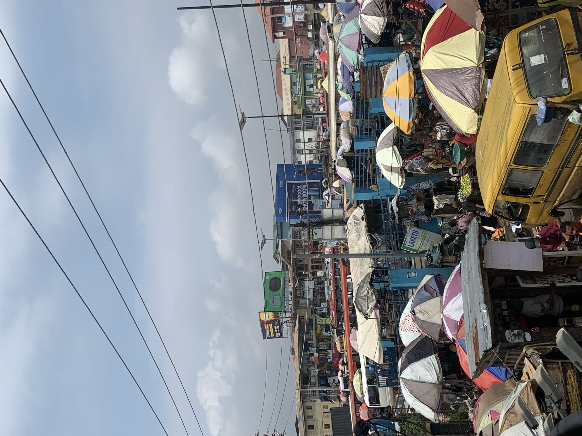 This is a photograph taken in Ikorodu, Nigeria, capturing a busy market scene under a partly cloudy sky. The foreground features a yellow minibus with its rear facing the camera. Several market stalls are visible, with shoppers and vendors actively engaged. Numerous umbrellas are erected over the stalls, providing shade. In the background, buildings of varying heights and architectural styles are present, along with more market activity and parked vehicles. Overhead power lines crisscross the sky. A prominent billboard for "RINOCOOL SHOWROOM" is visible in the mid-ground. The overall impression is of a bustling, vibrant urban marketplace.