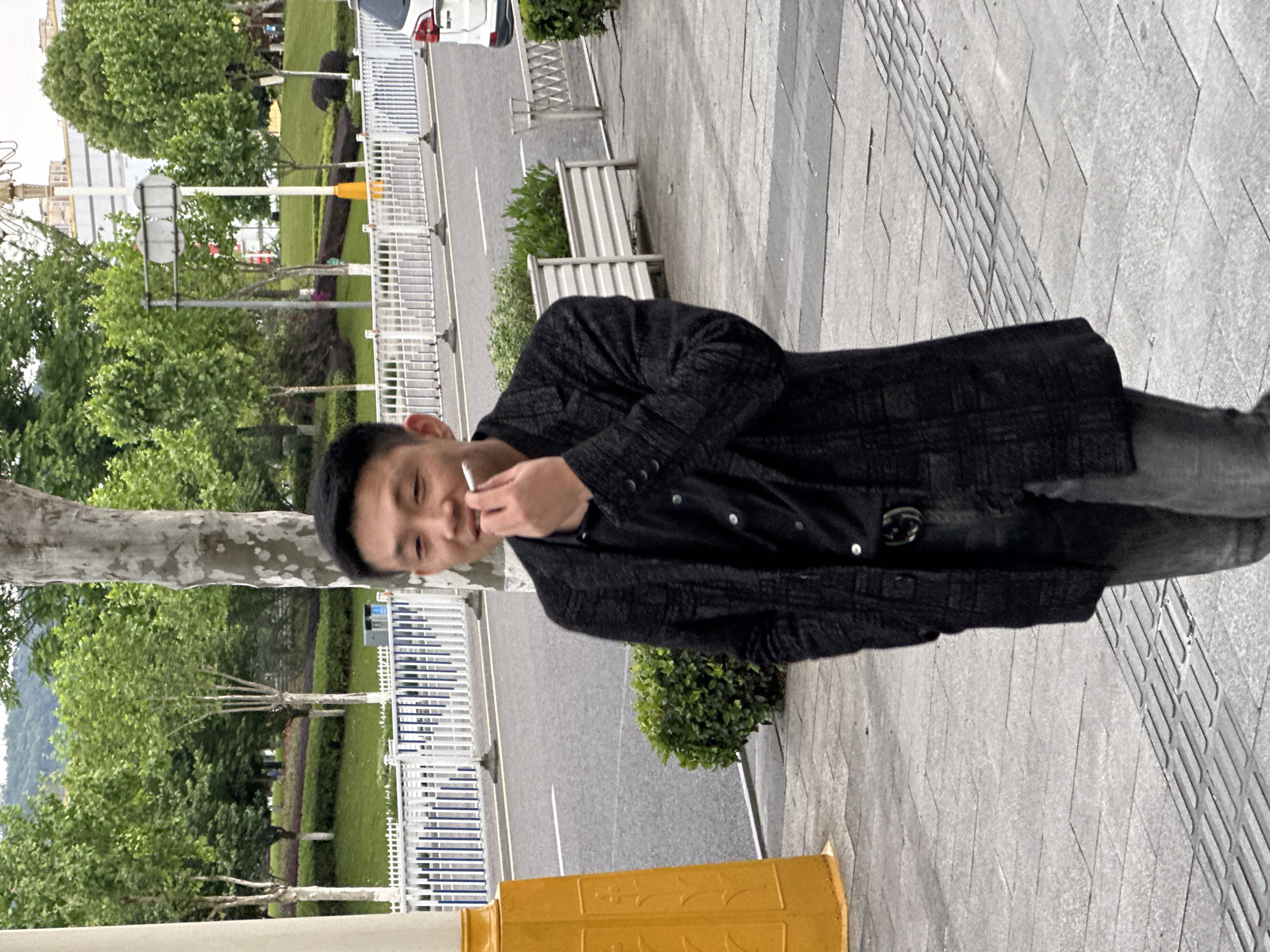 The image features a young East Asian man standing outdoors on what appears to be a paved sidewalk, under an overcast sky during daylight hours. He is seen from the waist up, facing slightly towards the viewer with a relaxed expression, possibly a faint smile or smirk. He holds a cigarette to his mouth, with a wisp of smoke suggesting he is in the process of smoking.
The man is dressed in a dark, subtly patterned long coat or jacket over a dark shirt with several light-colored buttons. A dark belt with a silver-toned buckle is visible at his waist. He has short, dark hair.
In the foreground, to his left, part of a tree trunk is visible, and a bright yellow street barrier or bollard occupies the lower left corner of the frame. The sidewalk he stands on is composed of various shades of grey, rectangular, and textured paving stones. Behind him, across a green area with bushes and trees, runs an asphalt road with a distinct white lane marking.
The background further reveals more greenery, including trees and bushes, a white metal fence, and some street furniture. Distant hills and urban structures are visible under the muted light of the sky. A white car is parked on the far right side of the road. The setting suggests an urban or suburban environment, possibly a park or green belt alongside a street in Jiangjin, China. No legible text is visible within the image.