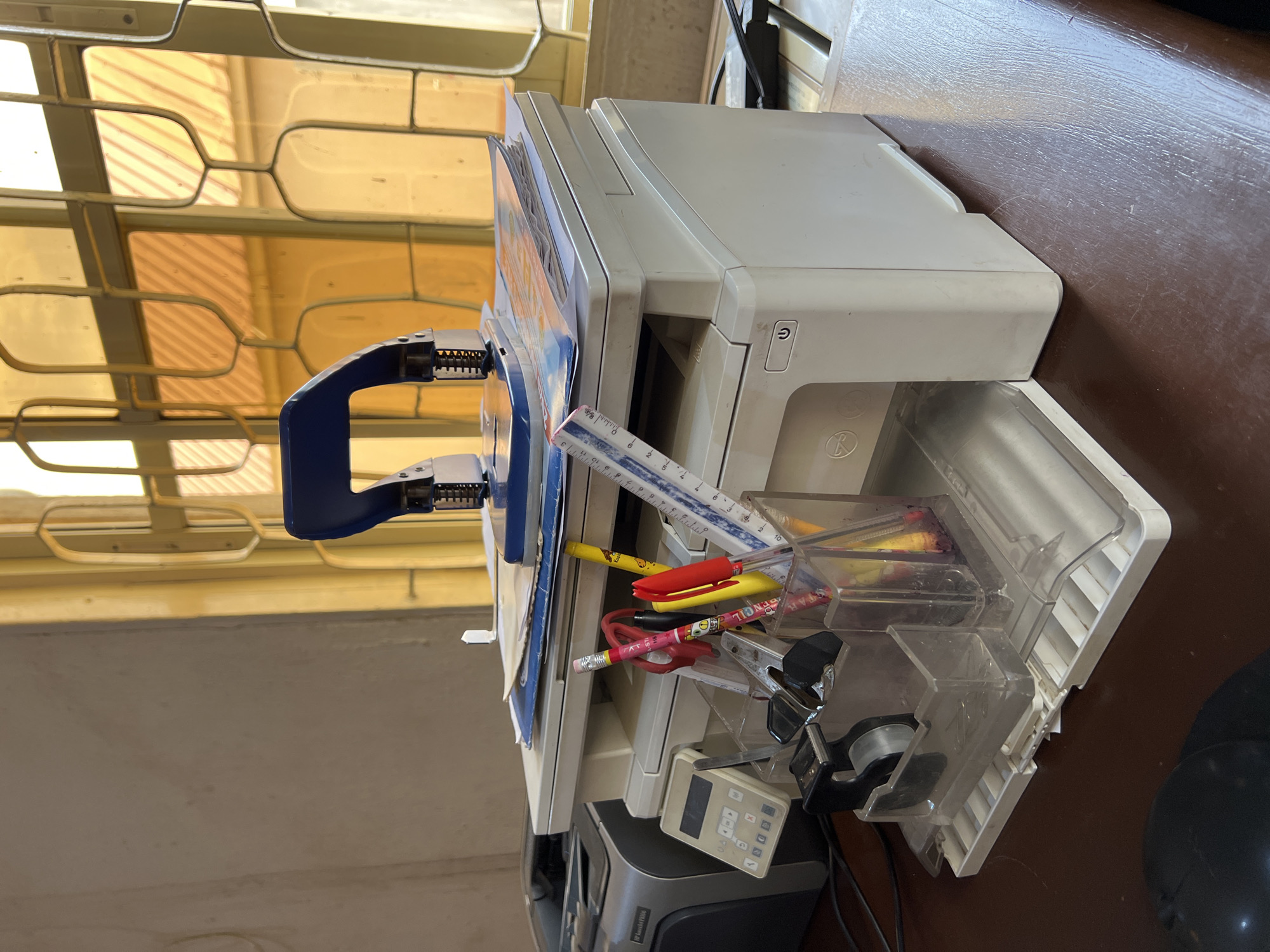 The image shows an office scene with a printer on a desk. The printer has an open paper tray filled with various office supplies including pens, pencils, a ruler, and paper clips. A blue stapler rests on top of the printer. A calculator is positioned below the printer near its base. The desk is brown. A window with a patterned frame is visible in the background, providing natural light. The location is assumed to be in Jos, Nigeria. No people are visible.