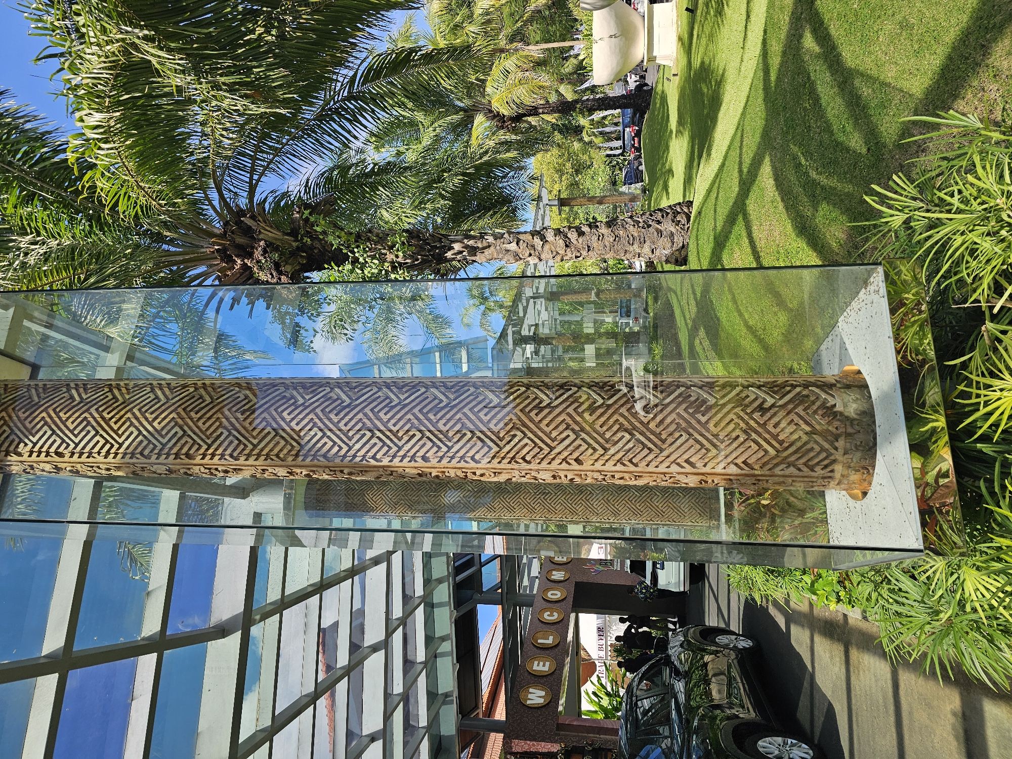 The image shows a building in Kuta Selatan, Indonesia. The building has a glass facade with metal support beams. A decorative wooden panel with geometric patterns is visible behind the glass. A "WELCOME" sign is positioned near the entrance. Outside, a black car is parked on the pavement. Lush greenery, including palm trees and plants, surrounds the building. The sky is clear and blue. Reflections of the surroundings are visible on the glass surfaces.