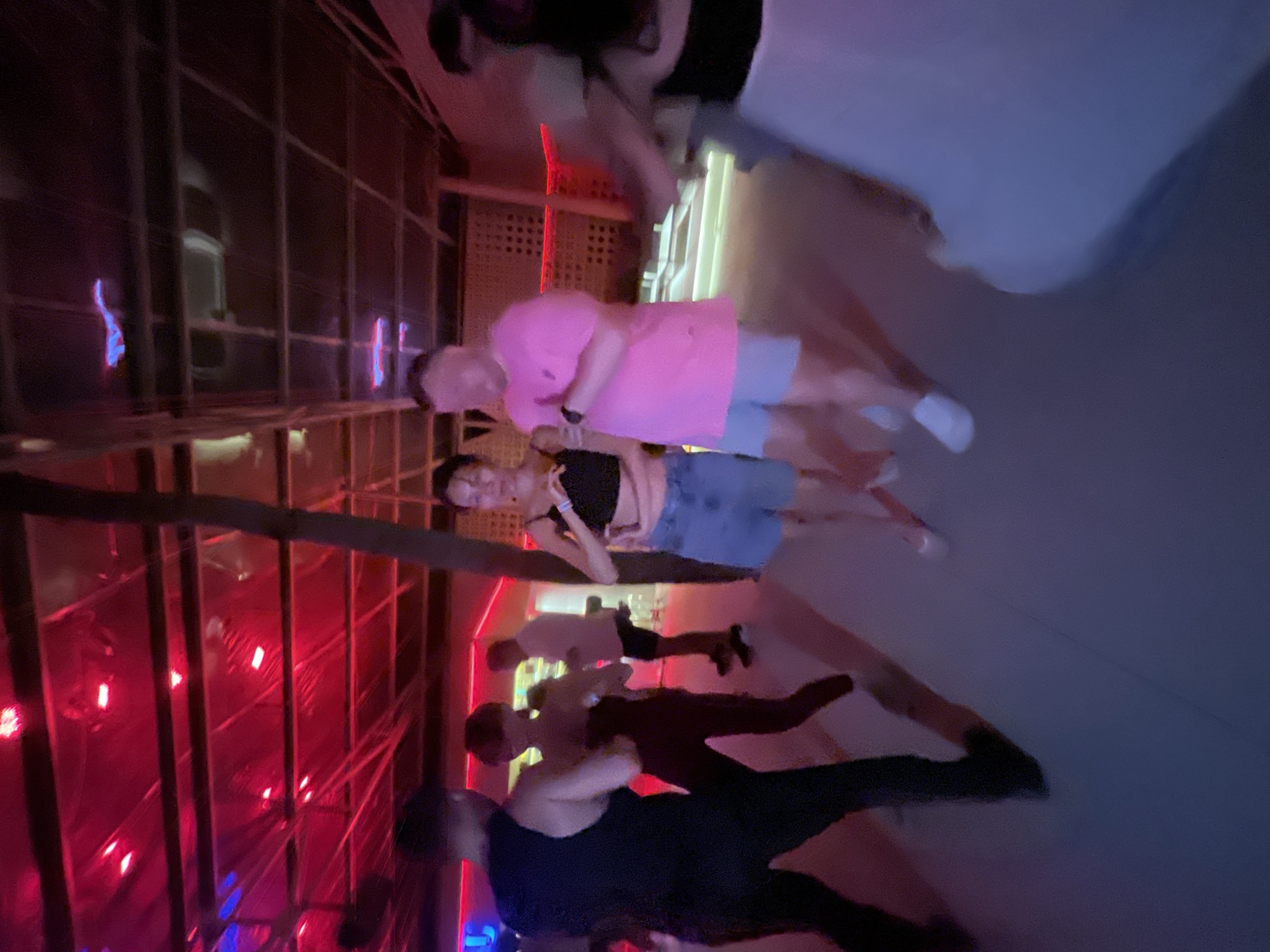 This media file shows a group of people in what appears to be a dimly lit, indoor venue with red and blue lighting. The image is oriented vertically, and the people are positioned throughout the frame. In the foreground, two individuals are visible, with one wearing a black tank top and the other a dark shirt. They seem to be engaged in an activity together, possibly dancing or interacting closely, with their bodies angled towards each other.
Behind them, another pair of people are more clearly visible. A person in a bright pink t-shirt and light-colored shorts is standing next to someone wearing a black top and denim shorts. They are also close to each other, suggesting a social gathering. Further in the background, more individuals are present, some of whom are partially obscured by the lighting and the structure of the venue.
The setting appears to be a space with a lattice-like structure or shelving unit, and there are red and blue lights illuminating the scene, creating a vibrant and possibly energetic atmosphere. This could be a nightclub, bar, or a party venue. The time of day is likely evening or night due to the artificial lighting and the general ambiance. The overall mood suggested by the image is one of socializing and enjoyment. While the specific event is not clear, it seems to be a casual social gathering or a night out. There is no visible text in the image. The location context provided is Da Nang, Vietnam.