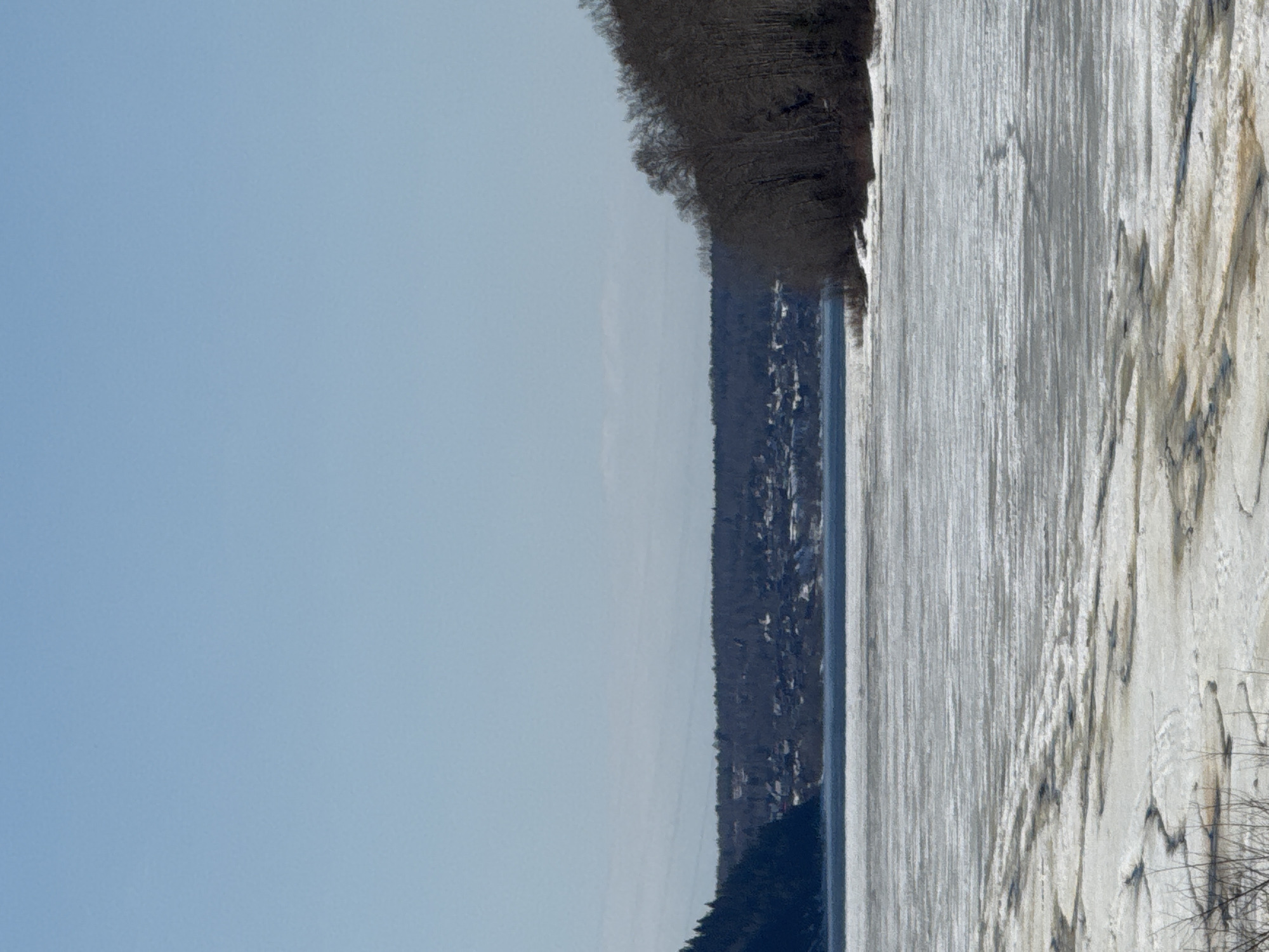 The image depicts a landscape with a clear blue sky covering the left half of the frame. On the right, a body of water, possibly a river or lake, is visible with ice covering its surface. The ice exhibits a textured pattern with varying shades of white and gray. A tree line borders the water, and a small town stretches along the distant bank. The image appears to be taken from a high vantage point, possibly a hill or building, and it's oriented sideways. Given the city indicated in the prompt, the image shows the Votkinsk Reservoir.
