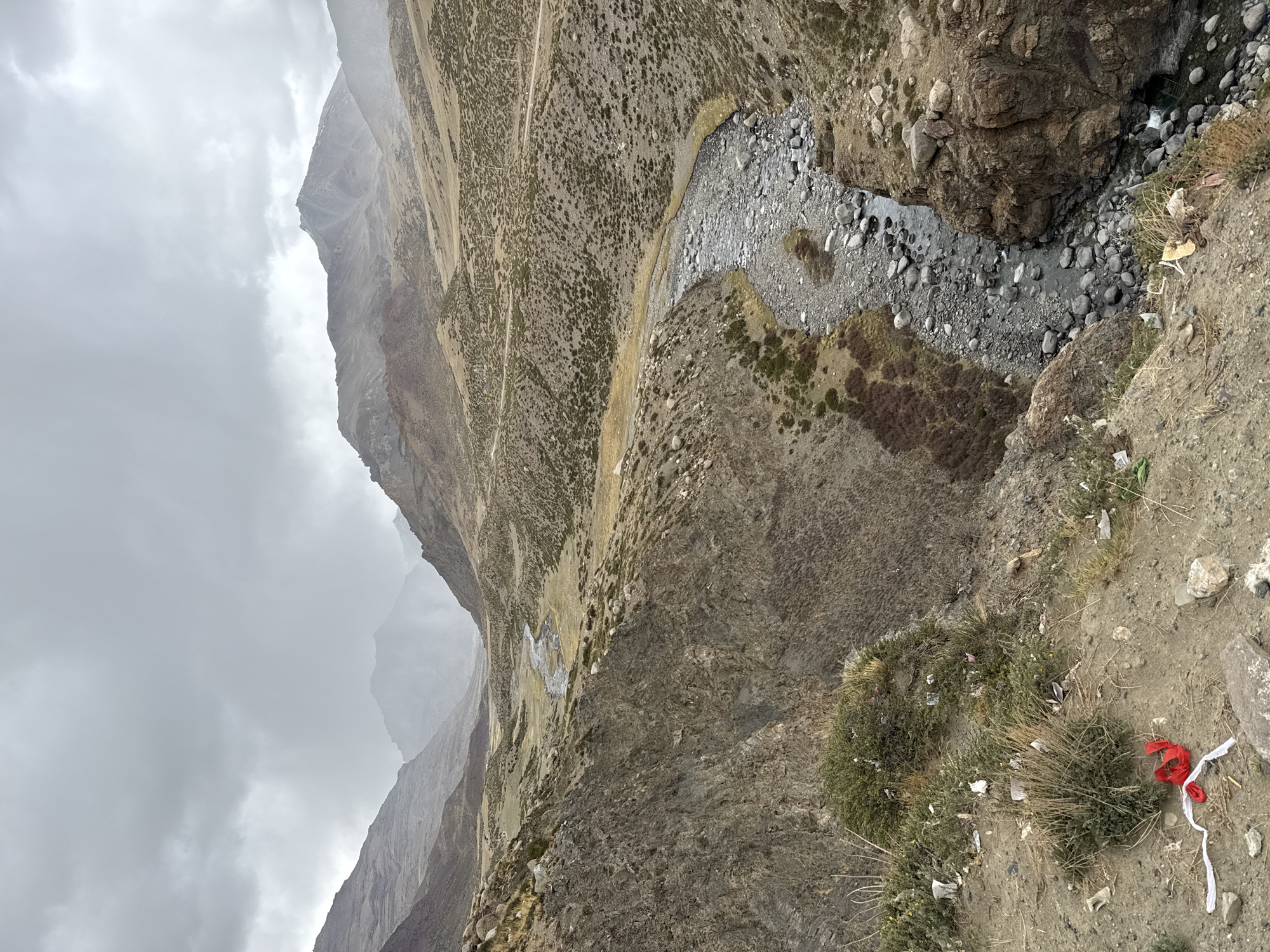 The image displays a vast and rugged mountainous valley under an overcast sky, captured from a high vantage point looking down into a riverbed and across to distant slopes.
In the immediate foreground, to the right, the ground is composed of dirt and rocks, with sparse patches of dry, bushy grass. Scattered among the vegetation are various pieces of litter, including white paper or plastic debris. Most prominently, a bundle of red and white fabric scraps is visible, tied together and lying on the ground.
Below this foreground, the valley floor reveals a narrow, rocky river or stream. Its bed is filled with numerous grey and white stones and pebbles of varying sizes, with shallow water flowing through. The riverbanks are steep and rocky, transitioning upwards into gradual slopes adorned with patchy scrub vegetation and scattered boulders. A faint, unpaved track or path can be observed winding along the mid-slope of the mountain on the opposite side of the valley.
The background is dominated by imposing, barren mountain peaks, their sharp contours partially obscured by the misty or cloudy conditions. The sky is entirely covered by a uniform layer of light grey clouds, indicating an overcast day with diffused lighting.
There are no people or animals visible, and no specific activity is taking place within the scene. No discernible text is present in the image. The landscape suggests a remote, high-altitude environment typical of mountainous regions, consistent with the context of Parga town, China.