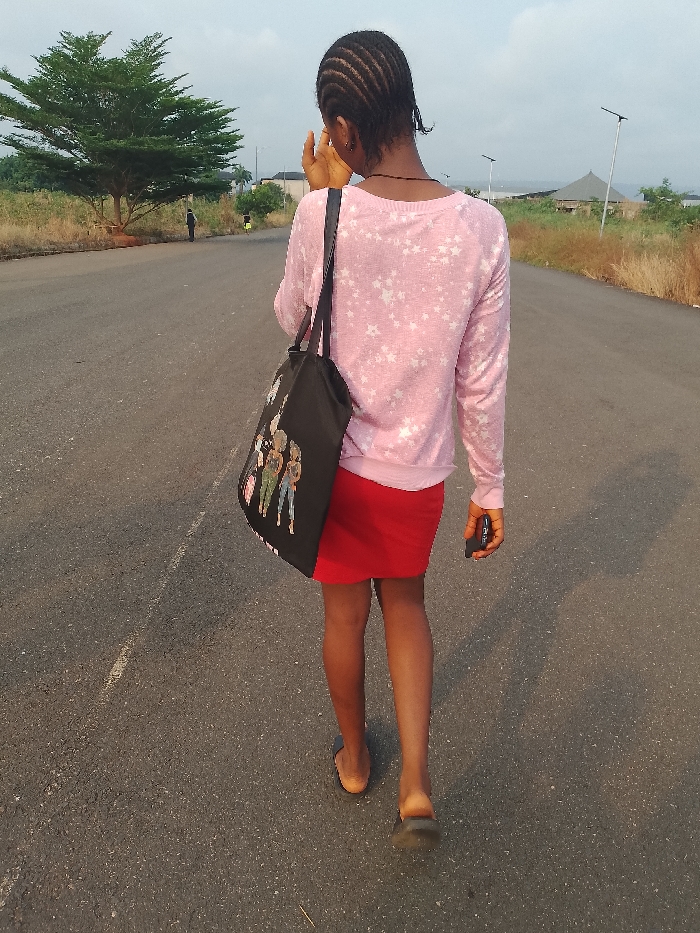 A young, slender woman with braided hair is walking away from the camera on a paved road. She wears a light pink, star-patterned long-sleeve shirt and a short red skirt. She carries a black shoulder bag with a design of three figures, and holds a small, dark object in her right hand. The road is lined with dry grassy areas and sparsely vegetated land. In the distance, a large tree and buildings are visible. The location is Umueshi, Nigeria, based on the metadata. The sky is overcast, suggesting an early morning or late afternoon time of day.