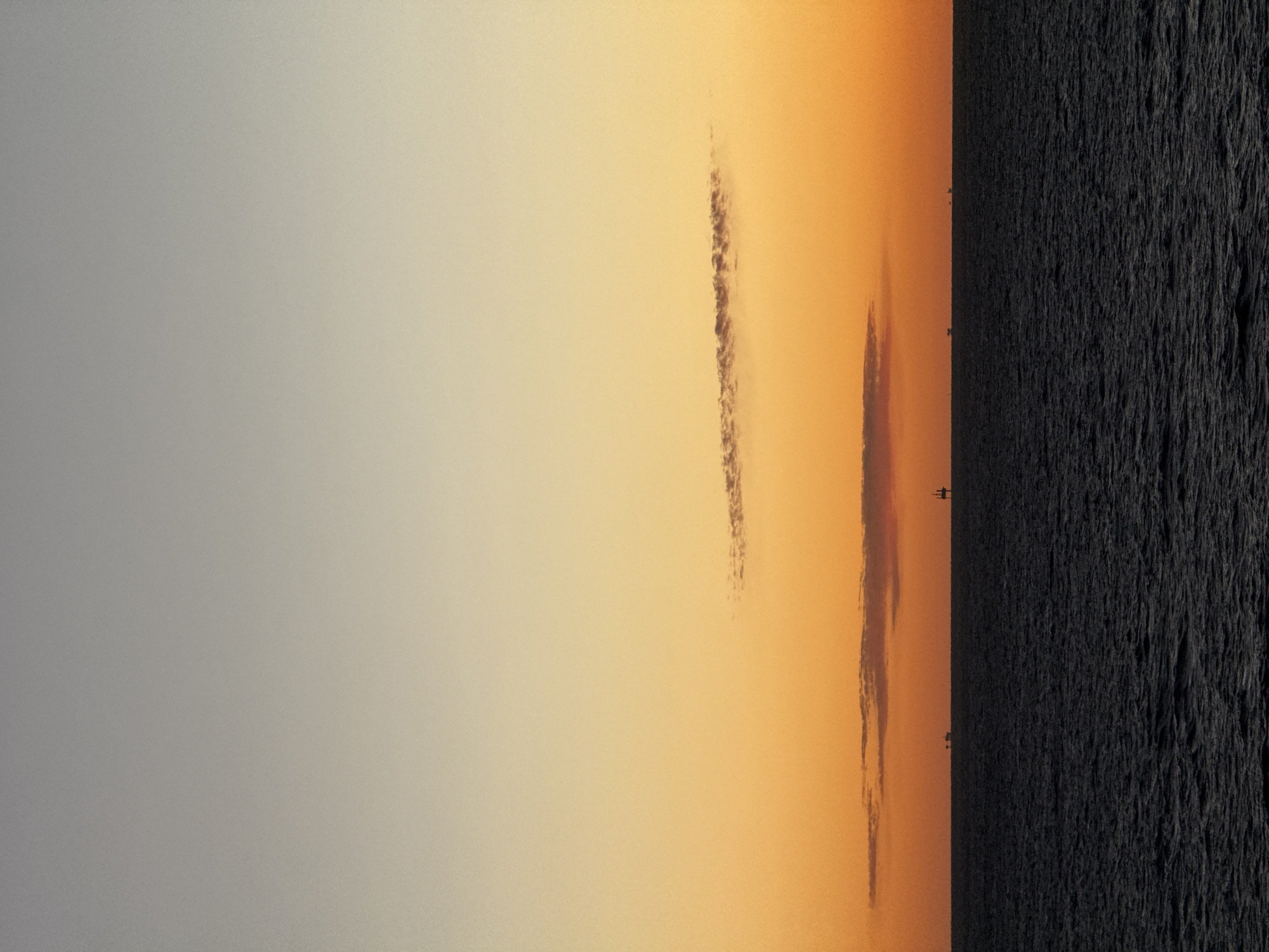 The image depicts a sunset scene over water, possibly an ocean or a large lake. The right side of the image shows a dark, textured surface, likely the water's surface with small ripples. The sky occupies the majority of the image. It transitions from a pale, gray-blue color on the left to a warm, orange-yellow hue towards the right. There are a couple of elongated, narrow cloud formations in the center of the frame. These clouds appear vertically oriented, adding depth to the sunset scene. In the distance, small, dark silhouettes appear on the horizon line between the water and the sky. The location and city are unknown based on the information provided.