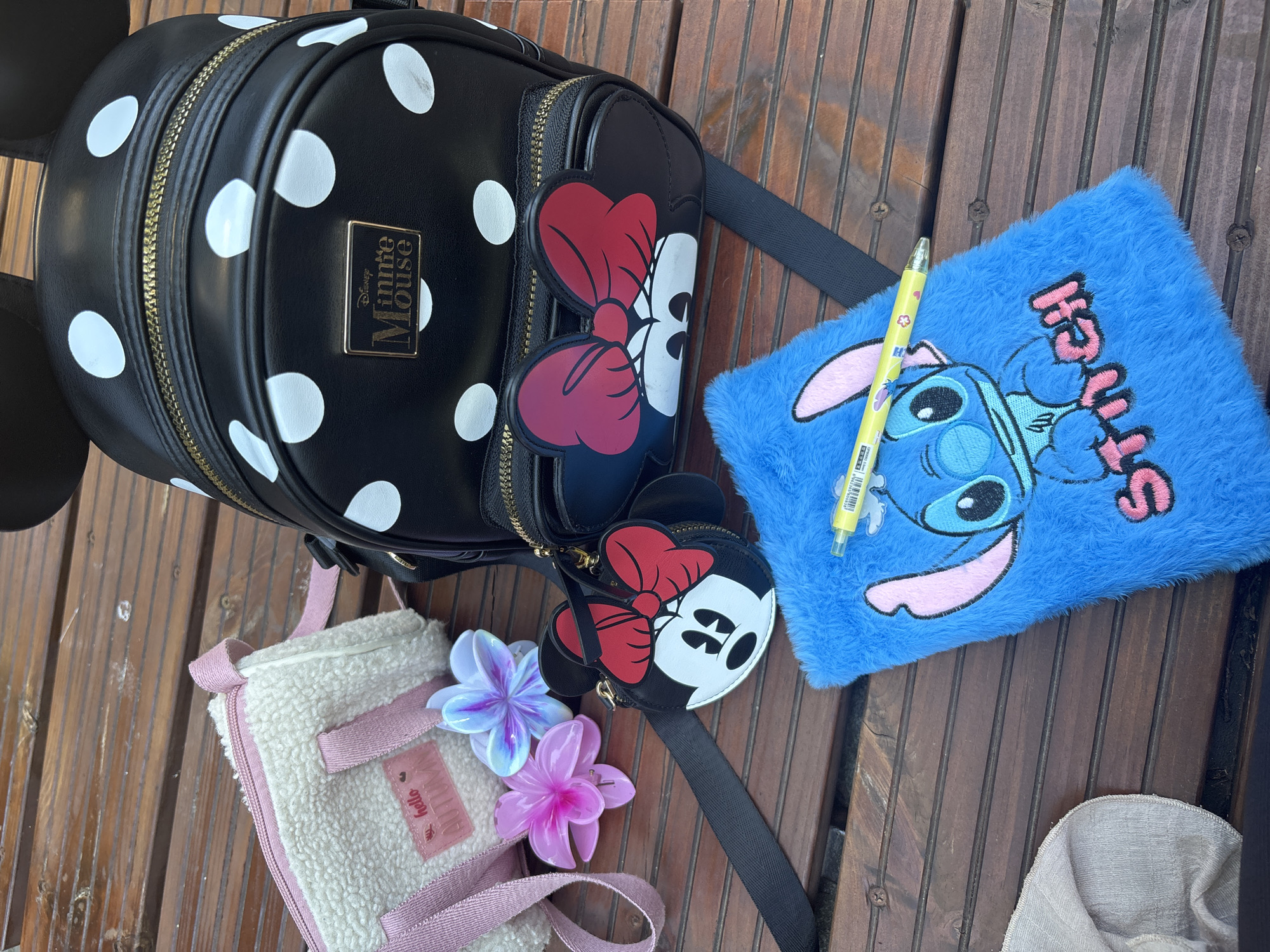The image displays a collection of Disney-themed items arranged on a wooden surface, likely an outdoor table. A black backpack with white polka dots and Minnie Mouse ears is prominently featured. Next to it, a small, round coin purse shaped like Minnie Mouse with a red bow is attached to a strap. A fuzzy, white shoulder bag with pink trim and a "hello AUTUMN" tag is positioned to the left. Two artificial flowers, one pink and one iridescent blue, lie between the shoulder bag and the Minnie Mouse coin purse. A bright blue, fuzzy notebook with a Stitch design is in the foreground, with a yellow pen resting on top. The wooden surface has visible grain and several screw heads. The background is blurred but suggests an outdoor setting. There are no people present. The scene is well-lit, with natural light casting soft shadows. The overall impression is of a curated display of merchandise.
