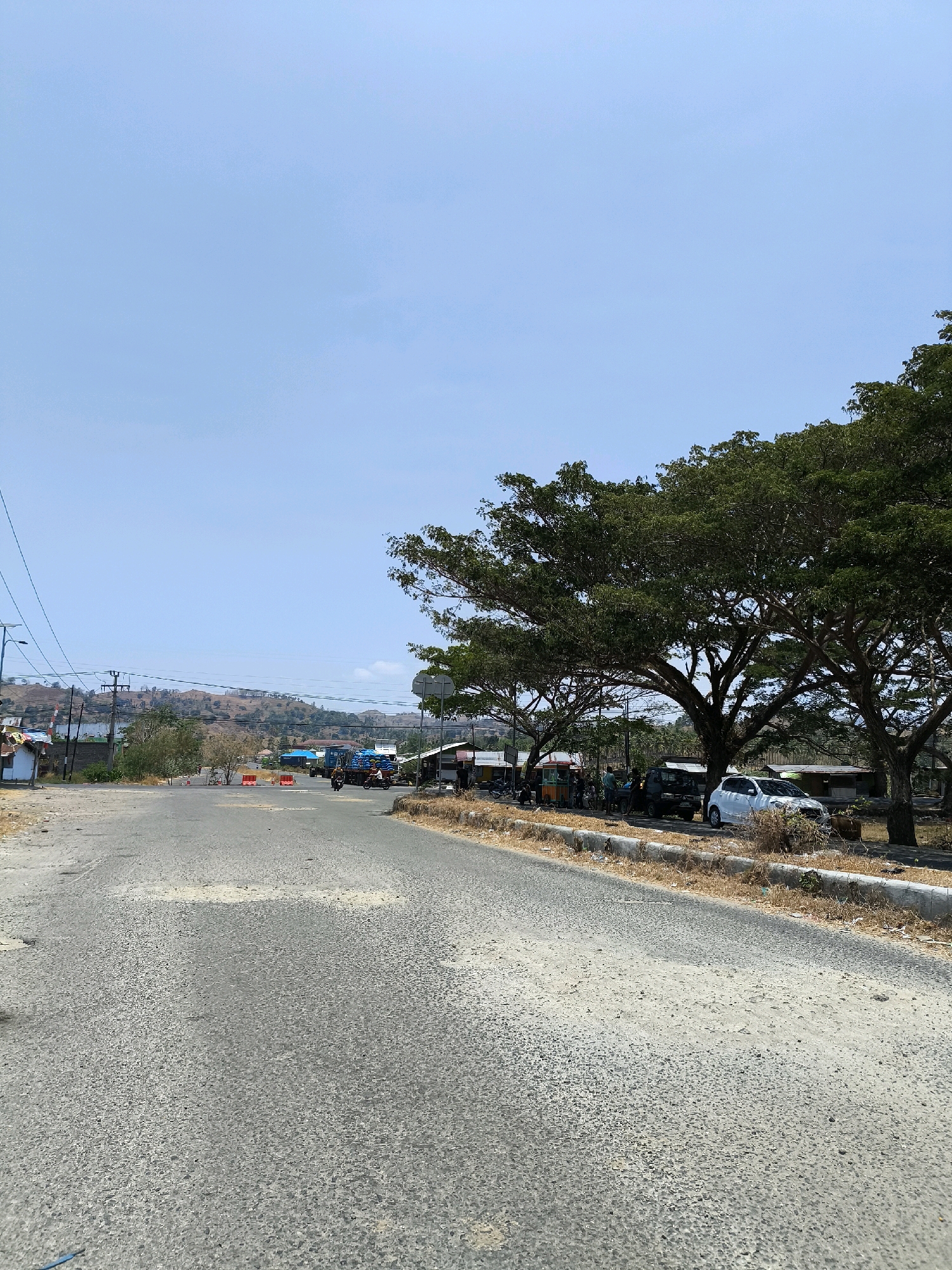 The image depicts a roadside scene in Kwandang, Indonesia, on a bright, clear day. In the foreground, a paved road with a rough surface shows signs of wear and tear, with patches of lighter-colored material and some visible potholes. To the right of the road, a concrete curb separates the pavement from a dry, grassy embankment. A line of large, mature trees with dense green foliage stretches along the embankment, providing shade.
Along the roadside, several small structures, possibly market stalls or small shops, are visible. A white car is parked under the trees, and further down the road, a dark-colored SUV and other vehicles are present. A truck with a blue cargo bed is also visible on the road. A few people are standing near the stalls and vehicles.
In the distance, the road continues towards a slight incline with hills covered in dry vegetation. Utility poles with wires are present on the left side of the road, alongside a few simple buildings and a small structure with a red and white flag. Orange traffic cones are placed on the road in the mid-ground, suggesting a possible obstruction or traffic control. The sky is a uniform pale blue, indicating fair weather conditions.