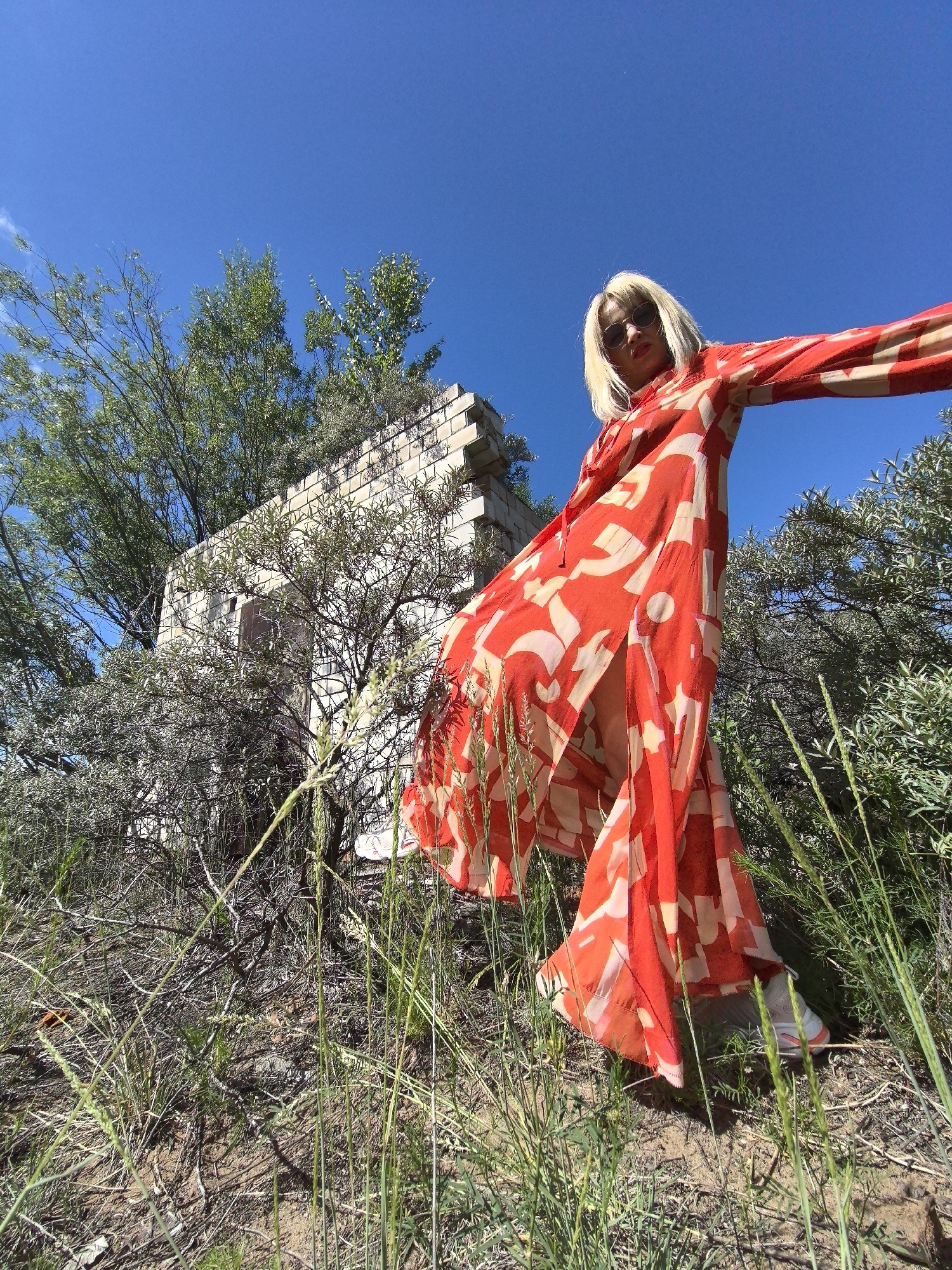 In an outdoor, low-angle shot, a person with blonde hair and sunglasses stands in front of a ruined brick structure in Noviy, Russia. The person is wearing a long, flowing red and white patterned dress and white sneakers. They are standing amidst dry grass and shrubbery with their arms outstretched. The sky is clear blue. The ruined structure appears to be made of light-colored bricks, with some of the top layers missing. Trees and more shrubbery surround the structure and the person. The overall scene suggests an abandoned or neglected outdoor location.
