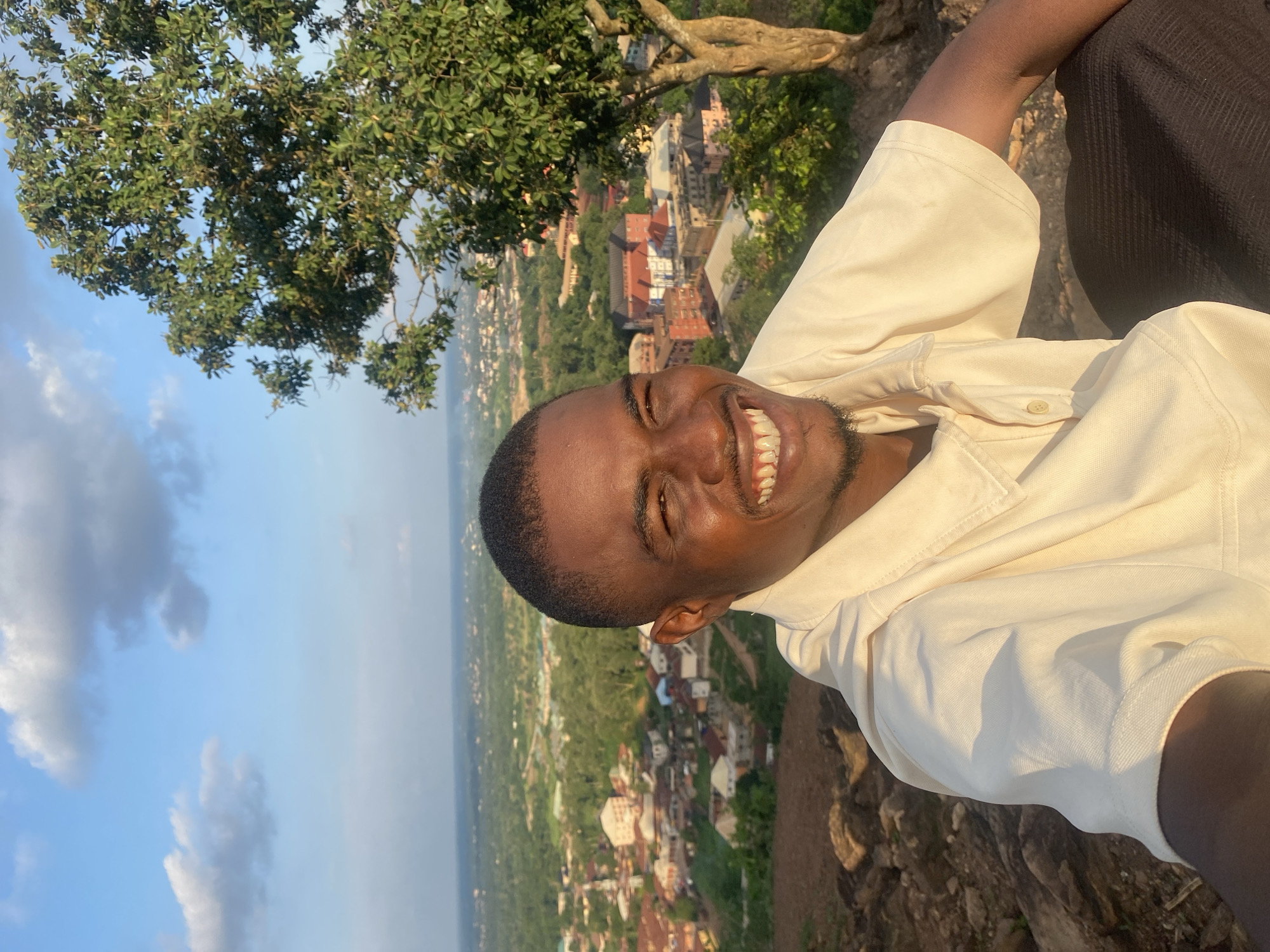 A close-up selfie captures a young Black man smiling broadly while sitting on a rocky hillside overlooking the city of Abakaliki, Nigeria. The man is positioned in the foreground, his face turned towards the camera and his body angled to the right. He is wearing a light-colored, collared shirt with short sleeves and dark trousers. Behind him, the city of Abakaliki unfolds in the distance, characterized by numerous buildings with red and white roofs nestled among lush green trees. A large tree with vibrant green leaves stands to the right of the man, partially obscuring the cityscape. The sky above is a clear blue with scattered white clouds, suggesting it is daytime, likely late afternoon given the warm lighting. The terrain in the immediate foreground consists of rough, brown rocks and sparse vegetation.