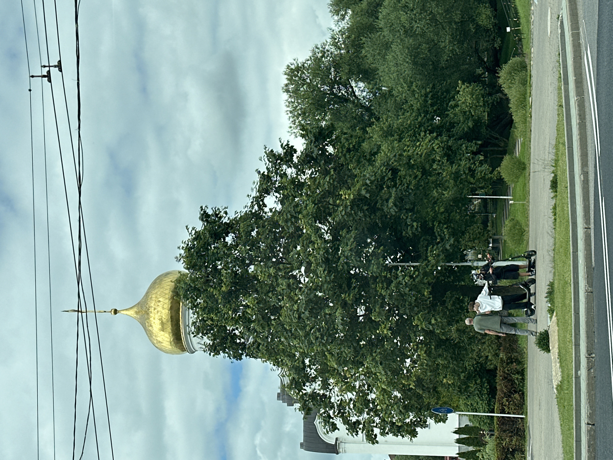 The image depicts a scene in Minsk, Belarus, featuring a prominent golden dome of a church against a cloudy sky. In the foreground, three individuals are gathered near electric scooters. An older man in a green t-shirt and gray pants stands to the left, facing away from the camera. Two younger individuals, one in a white hoodie and black pants, and another in a black outfit with a helmet, are positioned between the scooters. They appear to be interacting or preparing to ride.
Behind them, a large, lush green tree dominates the middle ground, partially obscuring the white facade of a building. The building appears to be a church or religious structure, with arched windows and architectural details visible. A blue circular sign with a bicycle symbol is visible on a white pole to the left of the tree.
The background consists of more trees and a grassy embankment, suggesting a park or public area. Power lines are strung across the top of the frame. The overall setting suggests an urban environment with a mix of historical architecture and green spaces, common in Minsk. The time of day appears to be daytime, with diffused light due to the overcast sky.