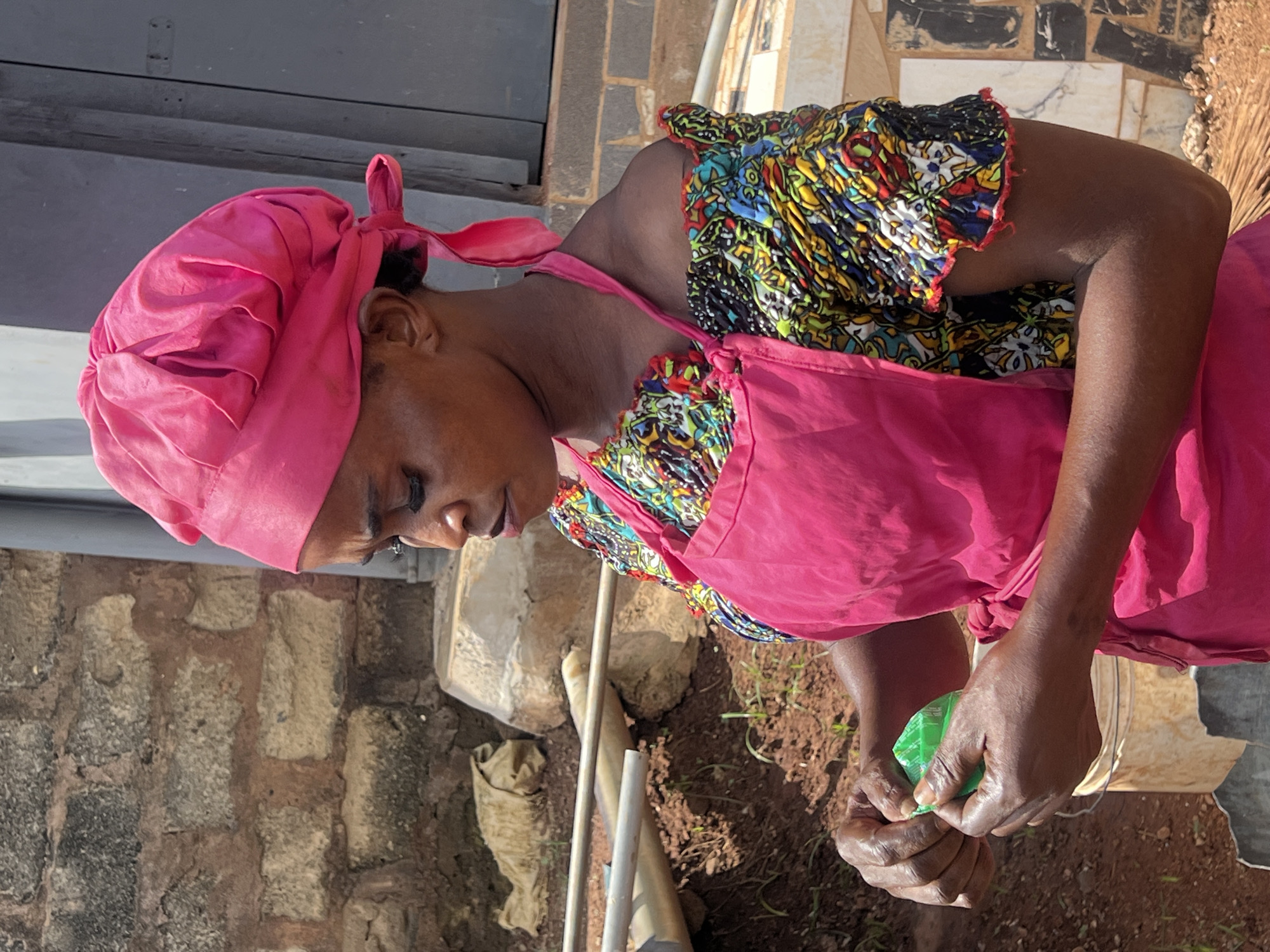 A woman wearing a pink headscarf and a bright pink apron over a vibrantly patterned off-the-shoulder top is captured in profile in Nkpor, Nigeria. Her dark skin is visible at her neck and arms, and her hands are engaged with a small green object, appearing to open or peel it. The background includes a brick wall, a large concrete structure, and a metal pipe. The ground is covered in reddish-brown soil with sparse vegetation. The lighting suggests it is daytime, with sunlight illuminating the scene.