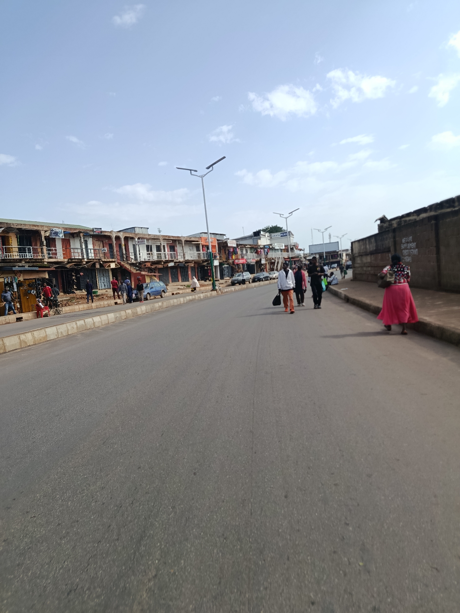 The image depicts a street scene in Jos, Nigeria, likely on a commercial street. On the left side of the street, there are multi-story buildings housing shops and residences. The buildings have a rustic, weathered appearance with balconies and open storefronts. Several people are visible on the sidewalk in front of these establishments, some appearing to be shopkeepers or customers. One person is with a bicycle. A blue car is parked near the shops.
The street itself is paved and appears to be a main thoroughfare. Several individuals are walking along the street, heading in different directions. One person in the foreground on the right is wearing a bright pink skirt and a patterned top. Further down the street, a group of people are walking together, some carrying bags.
The environment suggests an urban setting with elements of everyday commerce. The sky is a pale blue with scattered white clouds. Streetlights are visible along the side of the road, indicating it is a developed area. The overall atmosphere appears to be that of a moderately busy day in a Nigerian city.
