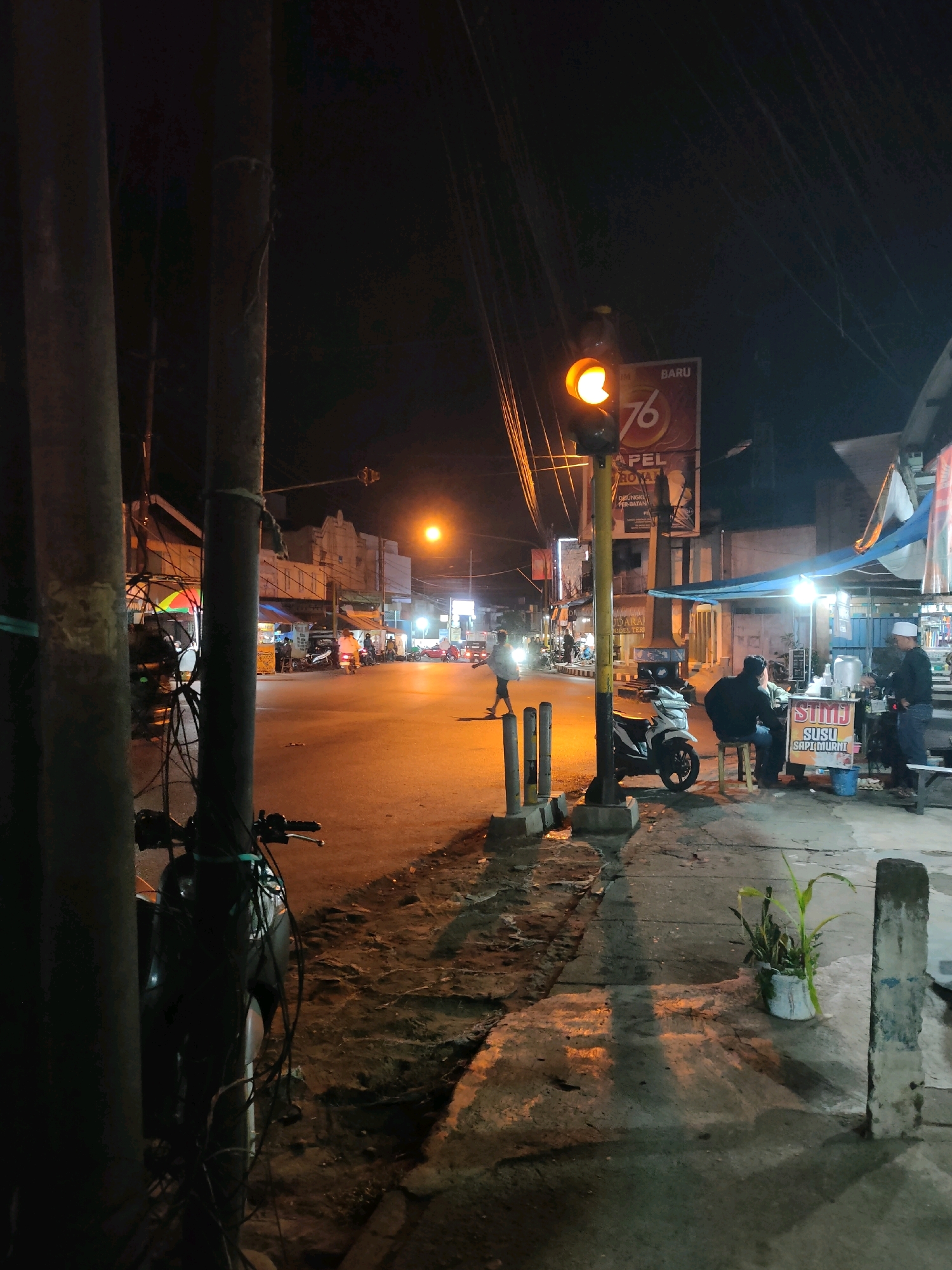 The image depicts a nighttime street scene in Codo, Indonesia. A traffic light is illuminated with a yellow signal, positioned at an intersection. In the foreground, a motorcycle is parked next to a thick utility pole. The street is paved and appears to be moderately busy with several vehicles, including scooters and cars, visible in the distance. On the right side of the street, a small food stall is set up under a blue awning, with two individuals seated and one standing, interacting with the vendor. A sign on the stall reads "STMJ SUSU SAPI MURNI," indicating it sells fresh cow's milk herbal beverages. The buildings lining the street are dimly lit, and various signs and posters are affixed to them. The overall atmosphere is one of a typical urban evening in Indonesia, with street lighting casting long shadows on the ground.