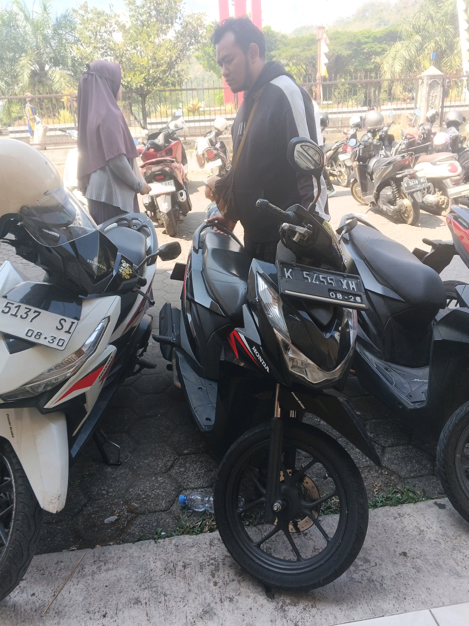 In Ungaran, Indonesia, on a sunny day, a man wearing a black and white hoodie stands next to a black Honda motorcycle with a license plate reading "K 5455 XH". A woman in a purple hijab and grey sweater stands to the left, facing away from the camera. Several other motorcycles are parked in the background. The scene appears to be a street or parking area, with palm trees and buildings visible in the distance. A plastic water bottle is on the ground near the motorcycles.