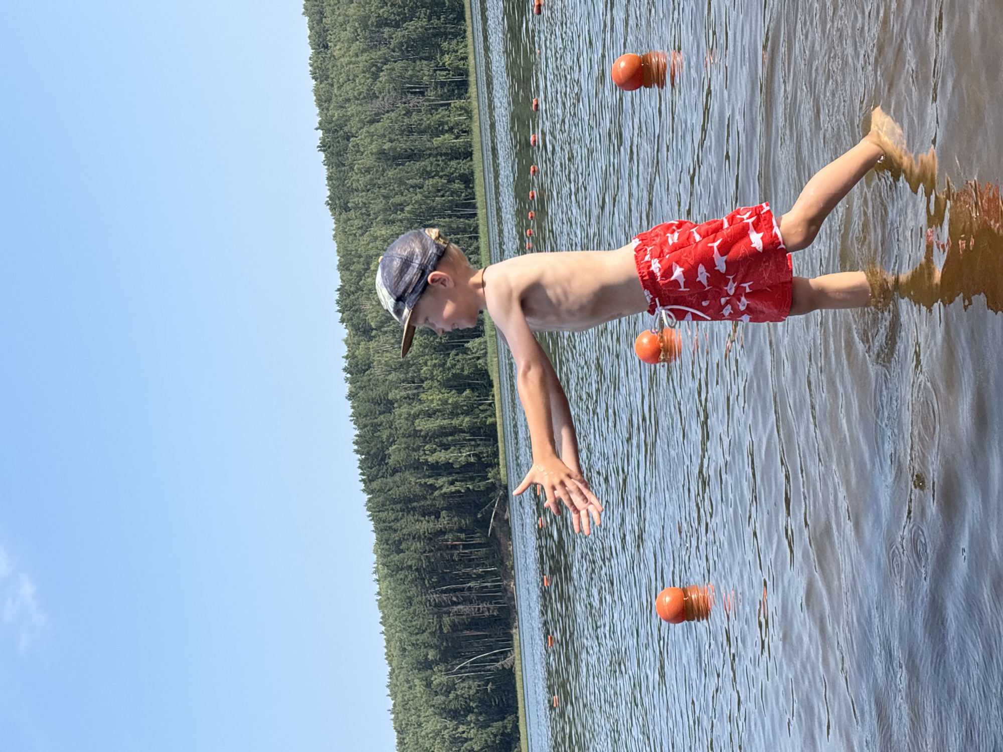 A young boy stands in shallow water at the edge of a lake, his arms extended forward as if reaching for something. He is wearing a baseball cap and red swim shorts with a white shark pattern. The water is calm with ripples, and several orange buoys are visible in the distance, marking a boundary on the lake. Behind the lake, a dense forest of green trees stretches across the horizon, under a clear blue sky. The scene appears to be a recreational area, possibly a beach or swimming spot, located in the city of Chaykovsky, Russia, known for its natural landscapes and proximity to the Kama River, which feeds into many lakes and reservoirs. The lighting suggests it is daytime, likely sunny.