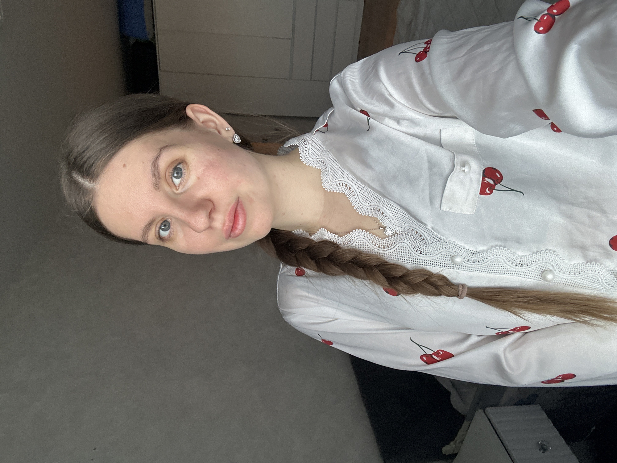 A young woman with long, light brown hair braided and pulled to one side is seen in a close-up shot. She wears a white collared shirt with red cherry print and a lace trim around the neckline. She is facing the camera with a slight tilt of her head, displaying a calm expression. The background appears to be a room, with gray walls and a white closet or cabinet. While the image doesn't provide enough detail to confirm the location, the query mentions Klin, Russia. Thus, the general environment is a residential interior, potentially located in Klin, Russia.