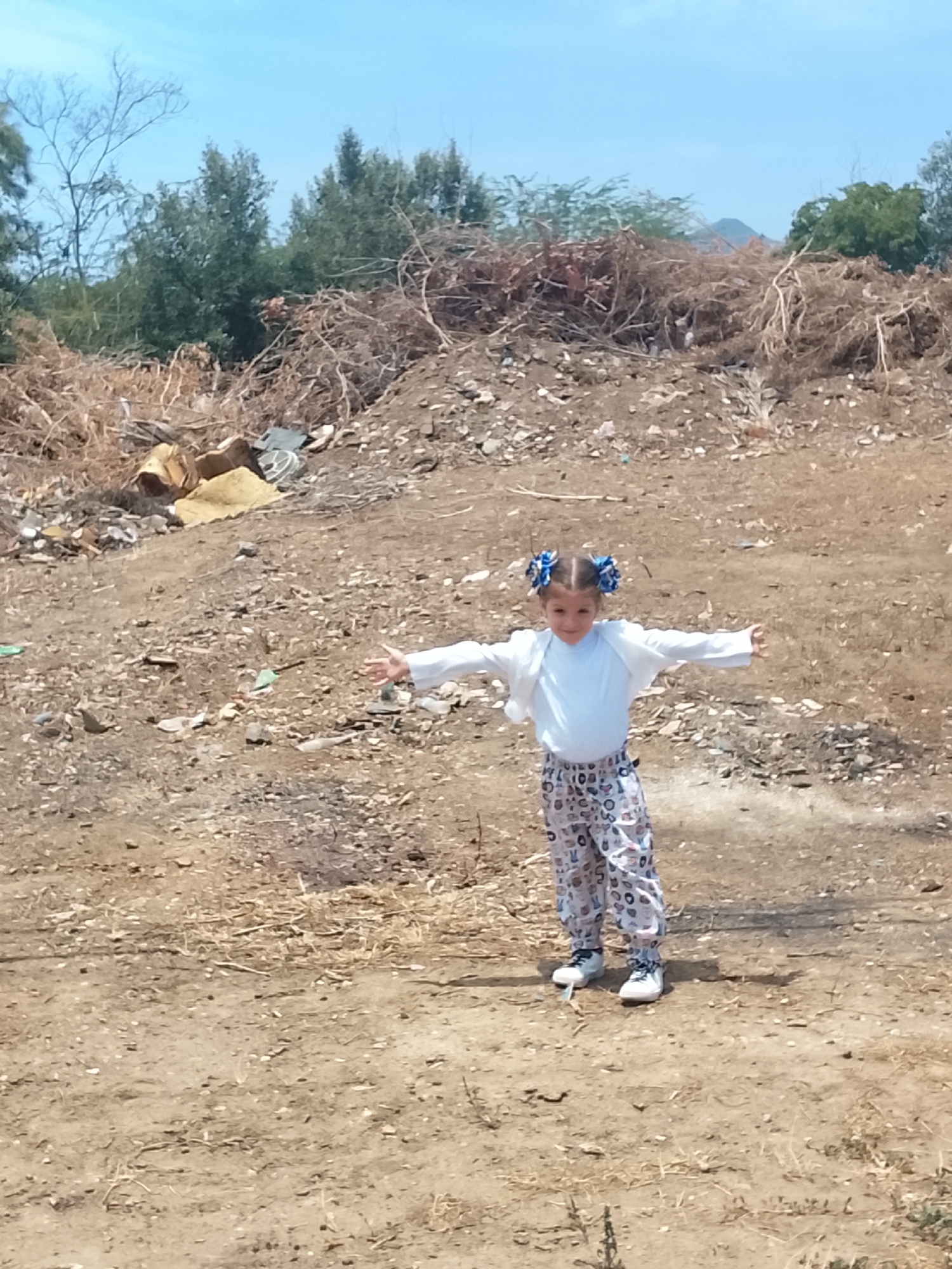 A young girl stands with arms outstretched in a barren landscape, possibly in Altagracia, Venezuela. She wears a light blue long-sleeved shirt, a white cardigan, patterned pants, and white sneakers. Her hair is styled in pigtails with blue ribbons. Behind her is a large pile of dry brush, scattered debris, and trash on dirt-covered ground. Some green trees and shrubbery are visible in the background, under a bright blue sky.