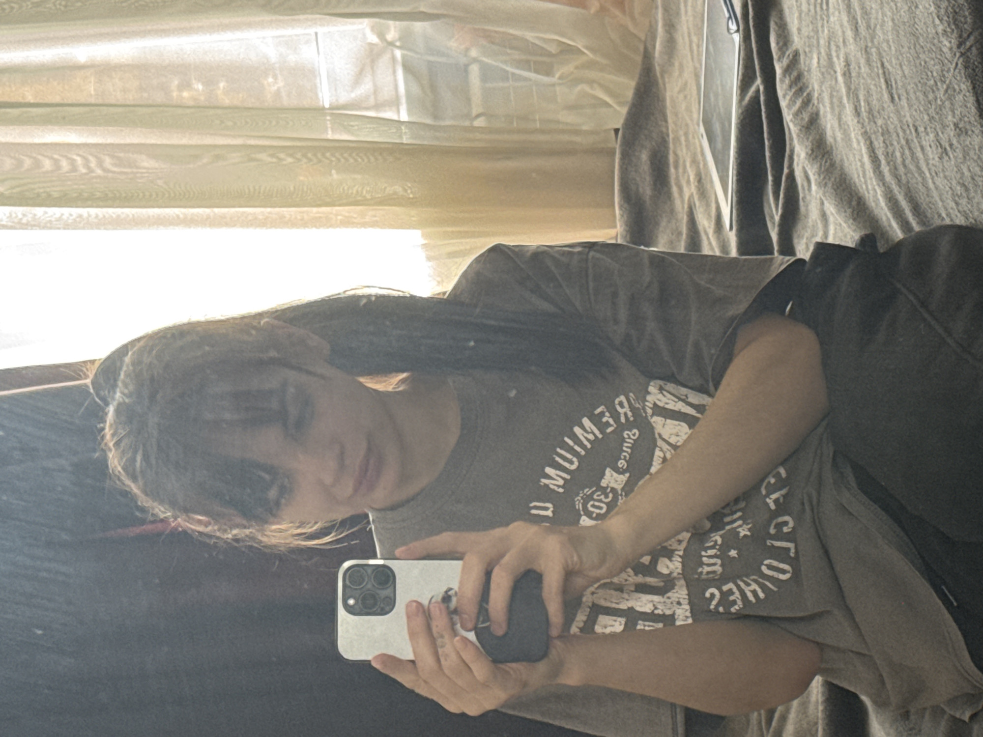 A person with long dark hair and bangs is taking a selfie in a mirror. They are wearing a gray t-shirt with a graphic print. The person is holding a white iPhone in their hands. The background includes a window with sheer curtains. There is a curtain to the right of the person. The location cannot be confirmed as Rostov-on-Don, Russia from the provided image.