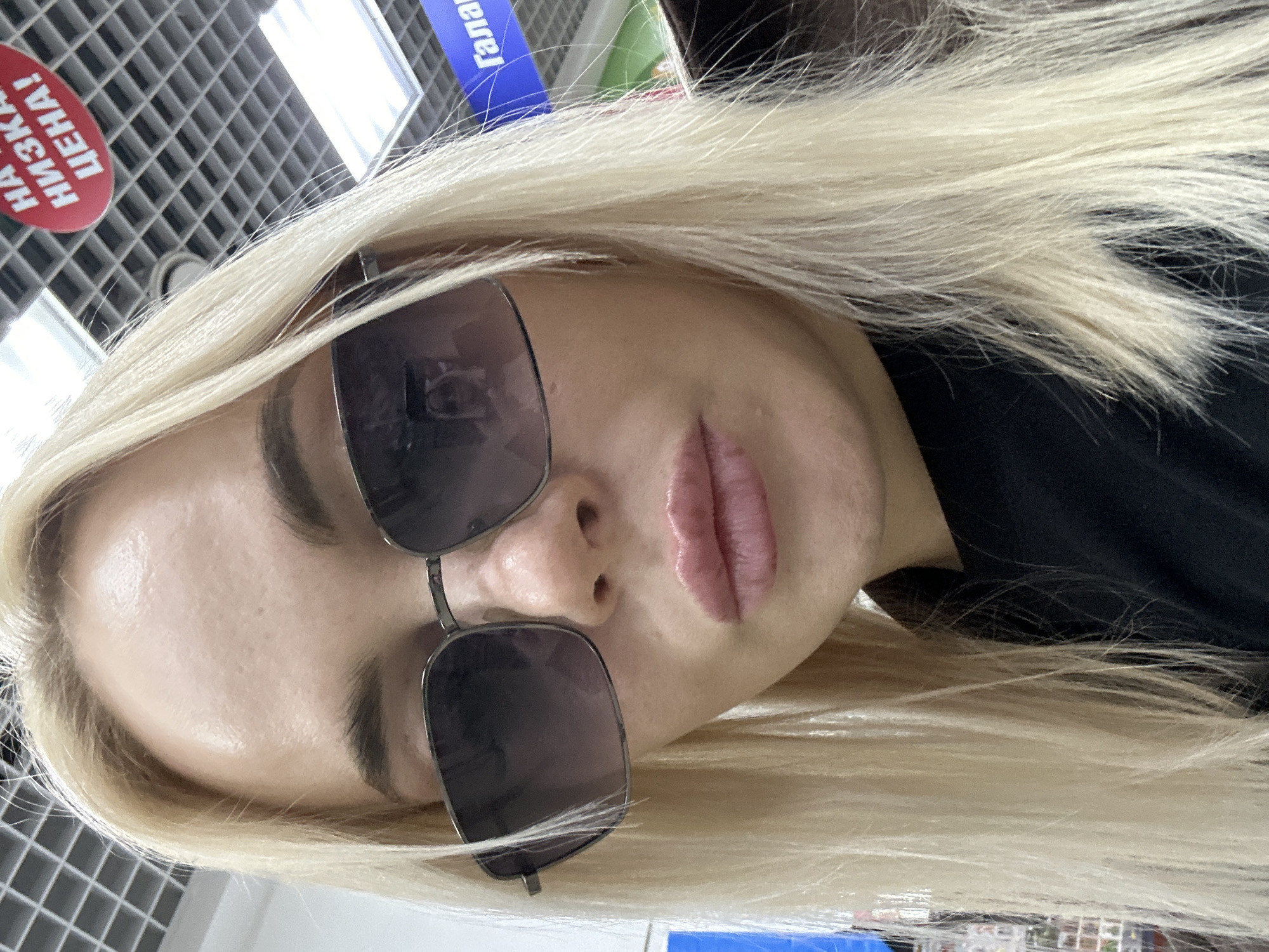 A close-up, slightly angled photo shows a woman with platinum blonde hair wearing dark-tinted sunglasses. Her lips are pink, and she has defined eyebrows. The background includes a ceiling with a grid-like pattern and various store signs in Russian. The photo was taken in the city of Megion, Russia.