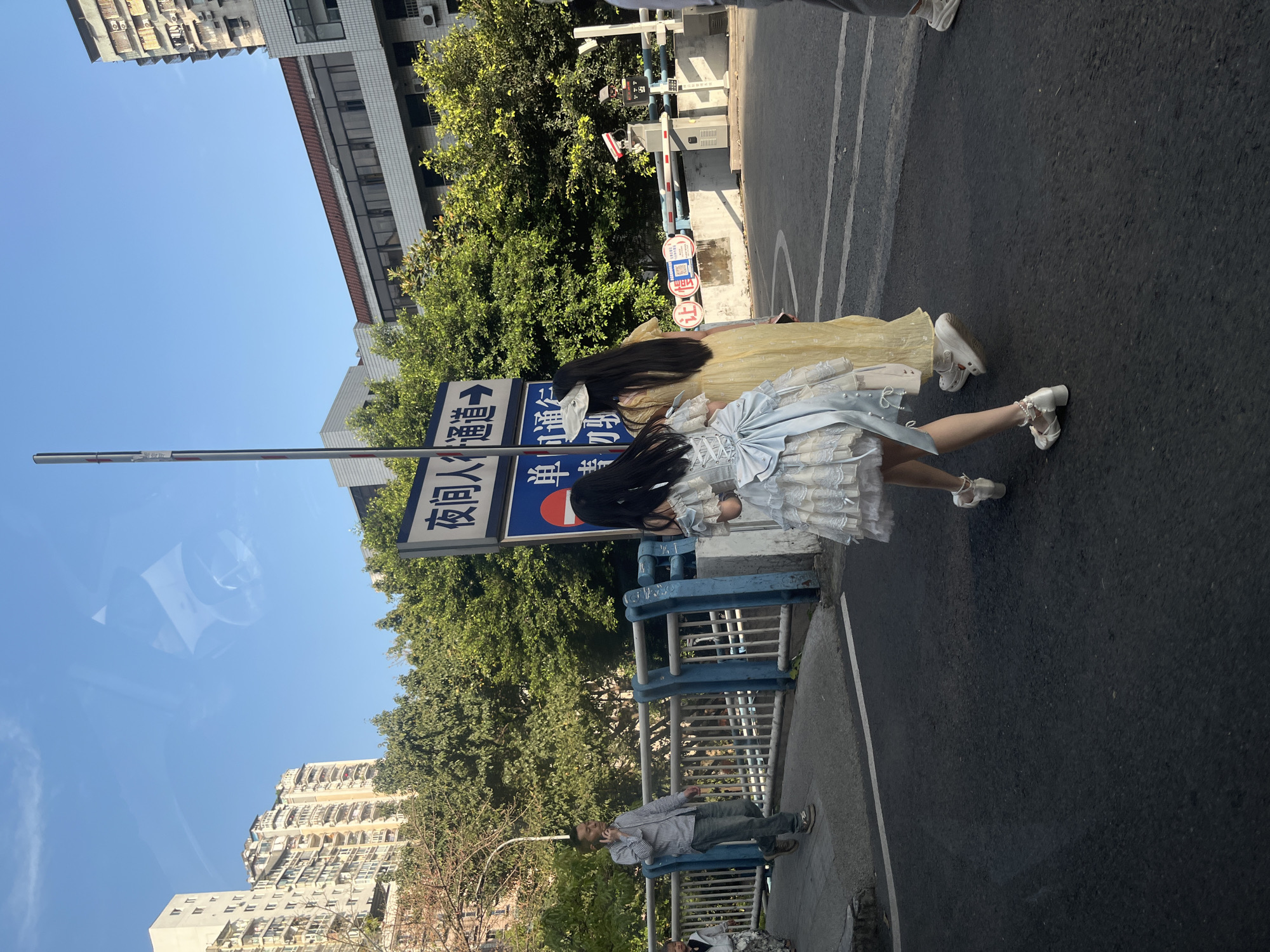 Two individuals in elaborate lolita-style dresses stand on the edge of an asphalt road, next to a sidewalk and a blue and grey metal railing. The person on the right wears a light blue or silver dress adorned with lace, ruffles, and a prominent bow, paired with white low-heeled shoes featuring pearl-like embellishments. The person next to them wears a yellow long dress. Both have long, dark hair.
Behind them, a tall pole displays blue rectangular street signs with white Chinese characters and arrows. One sign clearly reads "厦门道" (Xiamen Road), and below it "海滨路" (Haibin Road). A man in a striped shirt, jeans, and brown shoes stands on the sidewalk near the railing, with another person partially visible behind him.
Further back, a traffic barrier system with white and red arms, security cameras, and control boxes marks an access point. Green trees and foliage are abundant, particularly on the right side of the image. In the background, various residential and commercial buildings rise under a clear, bright blue sky, suggesting sunny weather during the day, likely midday or early afternoon given the harsh sunlight. On red circular signs near the barrier, the characters "禁" (forbidden/no entry) and "停" (stop/no parking) are visible. The scene depicts a street in what the provided context suggests is Liangjiang, China.