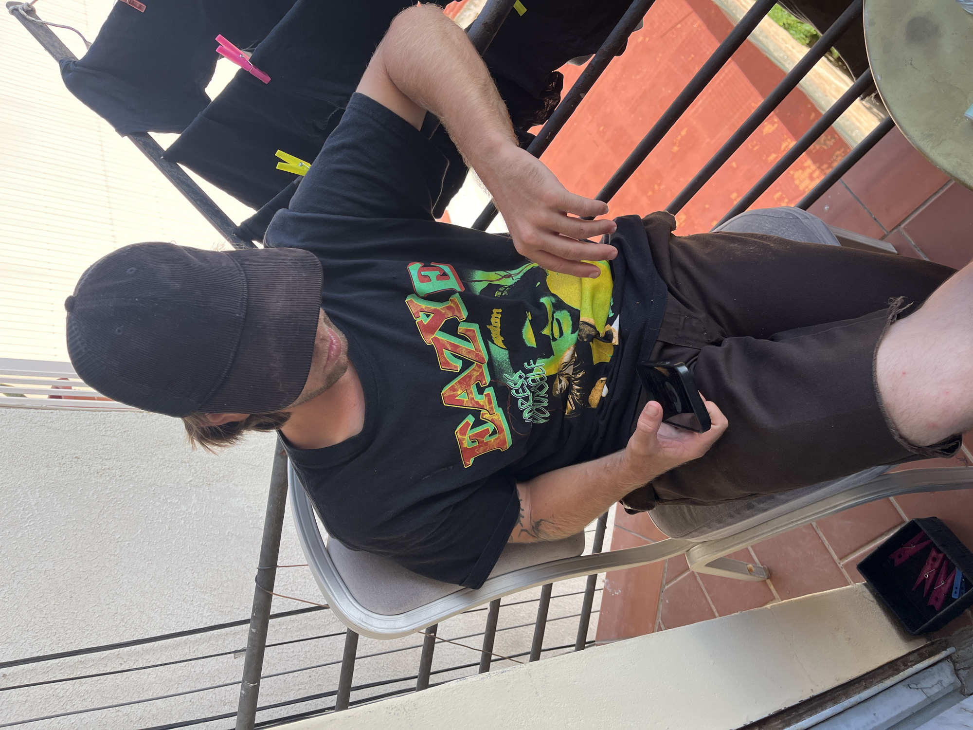 A man is depicted sitting on an outdoor balcony or patio, seemingly in a moment of leisure in Barcelona, Spain. He wears a black corduroy baseball cap, which largely obscures his face, and a black t-shirt featuring a prominent, colorful graphic print. The graphic includes large stylized letters in red and orange, which appear to spell "KAYI" or "KAYE," alongside an illustration and smaller, less legible text such as "DREES RU..." and "MOL." He is also dressed in dark brown shorts.
The man is seated on a light grey-framed chair with a padded seat, holding a black smartphone in his right hand and looking down at its screen. The scene is brightly lit by what appears to be daytime sun. The floor is tiled with reddish-brown terracotta. A black metal railing runs horizontally across the midground. To the man's left and slightly behind him, a black clothes drying rack holds dark garments secured by bright pink and yellow clothes pegs. On the tiled floor near his feet, a small black container holds additional pink and blue clothes pegs. A textured white wall is visible to the far left. The overall impression is of a casual, sunny day.