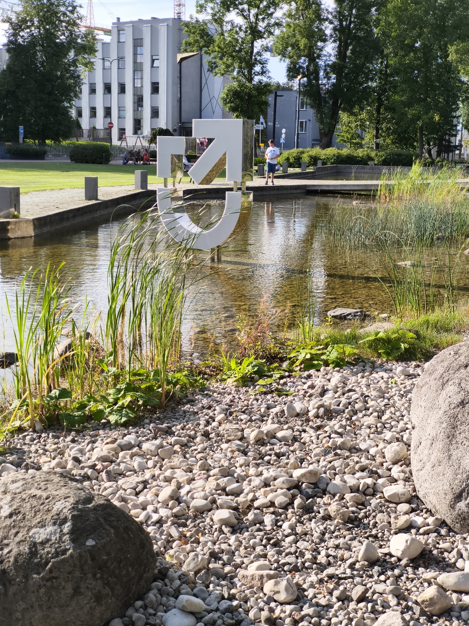 The image captures an outdoor scene with a pond in the foreground, surrounded by gravel and some large rocks. A large, white, abstract sculpture resembling a stylized male symbol is partially submerged in the pond. Tall reeds and green plants grow along the edge of the water.
In the background, a group of people, including several children, are gathered on a swing set on a grassy area. Further back, a modern, multi-story building with a light-colored facade and numerous windows is visible. Trees with green foliage are scattered throughout the scene. A young person in a white t-shirt and blue shorts is standing on a concrete walkway next to the pond. Two large construction cranes are also visible in the distance, suggesting ongoing development in the area.
This scene appears to be located in Tallinn, Estonia, likely in a public park or recreational area, given the presence of the sculpture, pond, playground, and modern architecture. The lighting suggests it is daytime, with sunlight casting reflections on the water and highlighting the textures of the gravel and rocks.